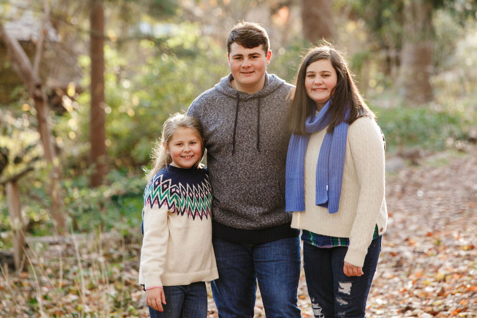 Three smiling people stand on a leaf-covered path in a wooded area at the Grange Estate. The younger two wear sweaters, and the older one wears a scarf, capturing a perfect family photo moment.
