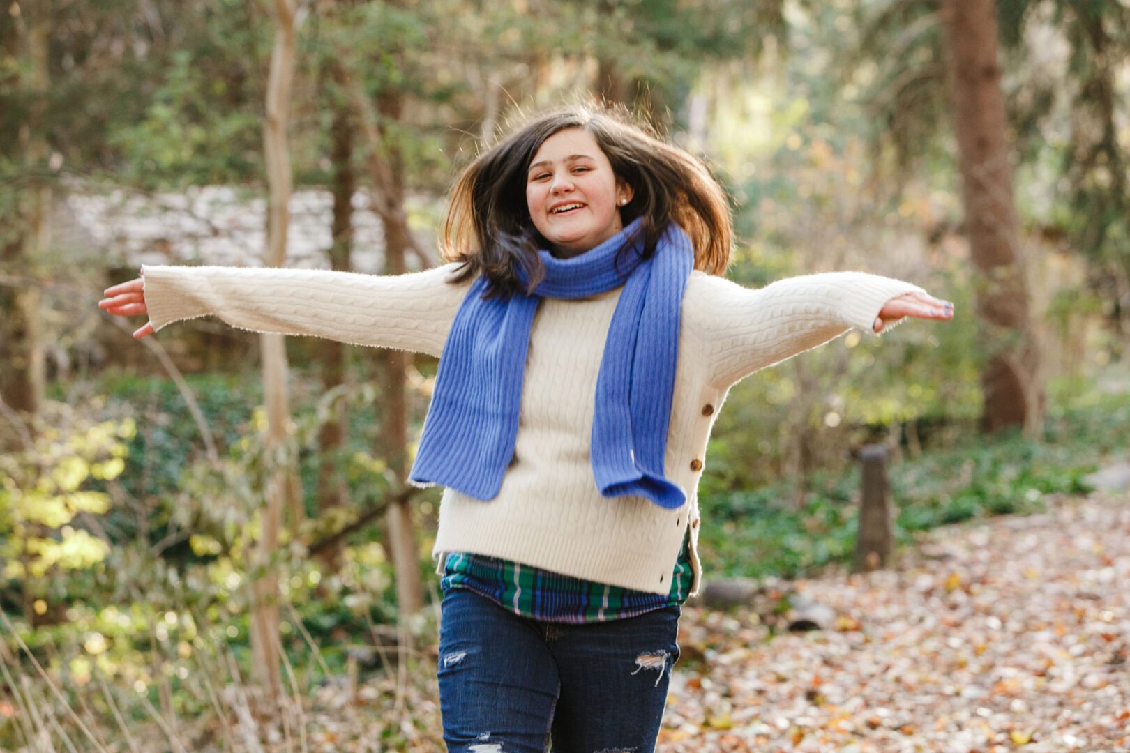 A person joyfully strolls through the Grange Estate, a wooded area covered with fallen leaves, wearing a white sweater, blue scarf, and jeans—perfect for family photos.