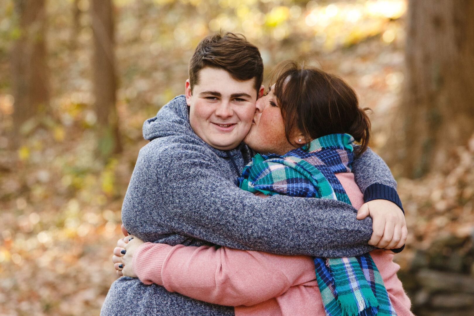 A woman wearing a scarf kisses a smiling boy on the cheek as they embrace in the wooded charm of Grange Estate, surrounded by fallen leaves.