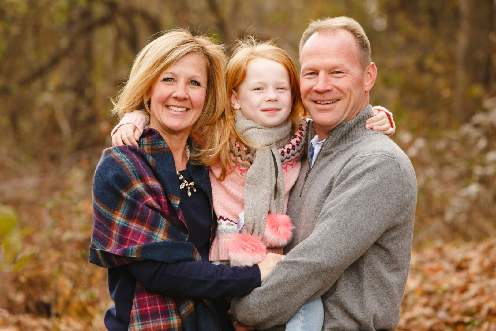 A smiling family of three poses outdoors for their Malvern Family Photos, with the child nestled between two adults in autumn clothing. The background features trees and fallen leaves, capturing a perfect seasonal moment.