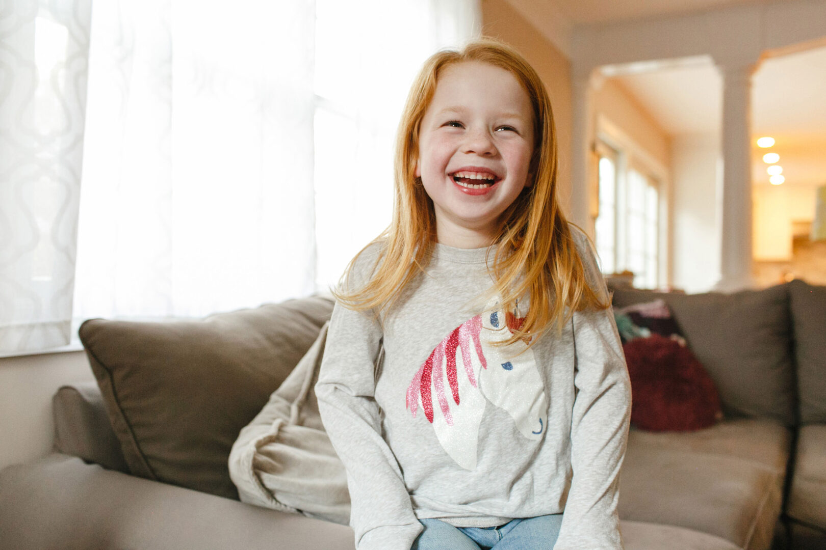 In a bright living room, a red-haired child sits on the couch, beaming in a gray unicorn shirt. This cheerful moment is part of the Malvern Family Photos series.