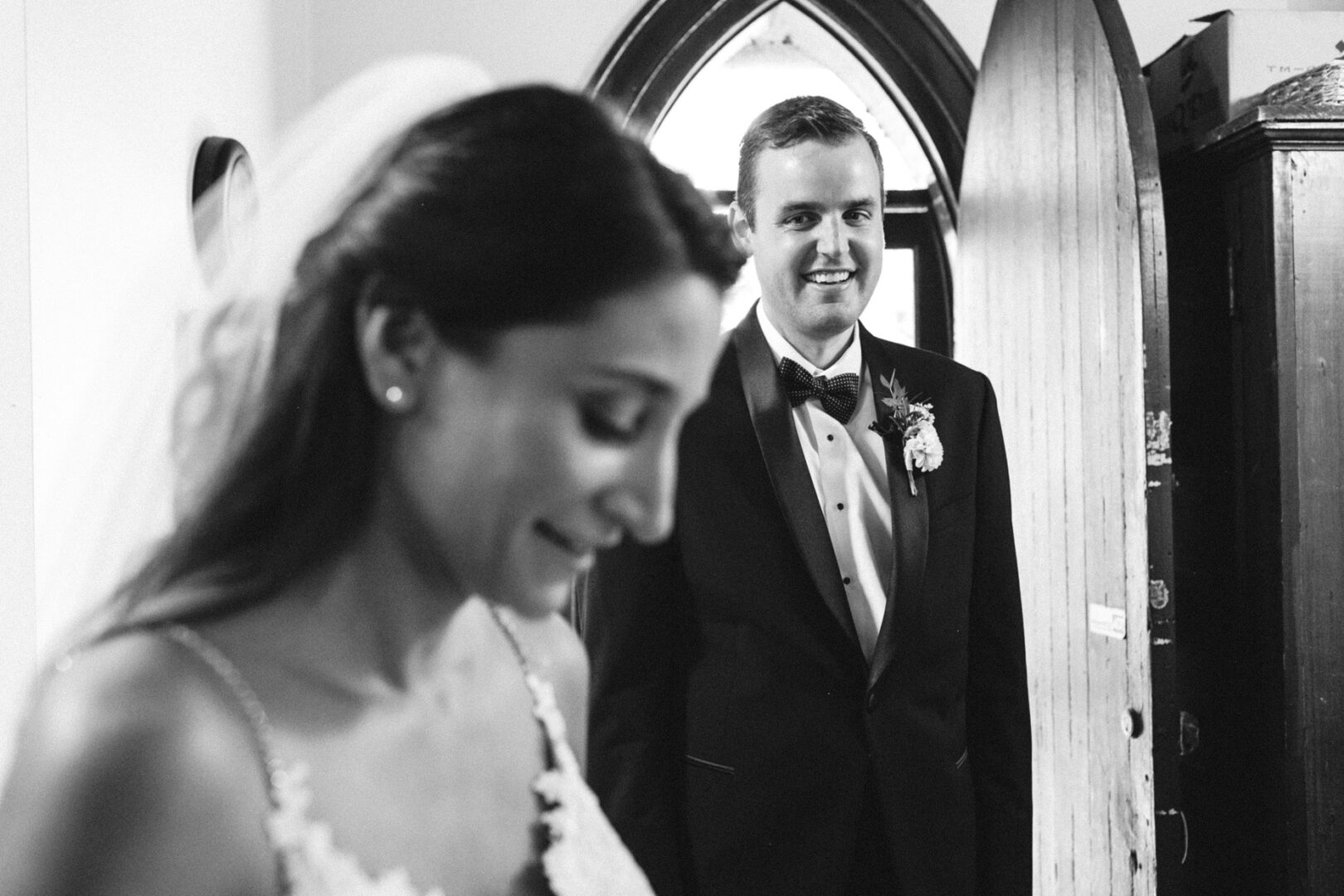 Bride and groom standing indoors, bride in foreground with veil, and groom in tuxedo smiling by the arched window at their Cape May wedding. Black and white photo.