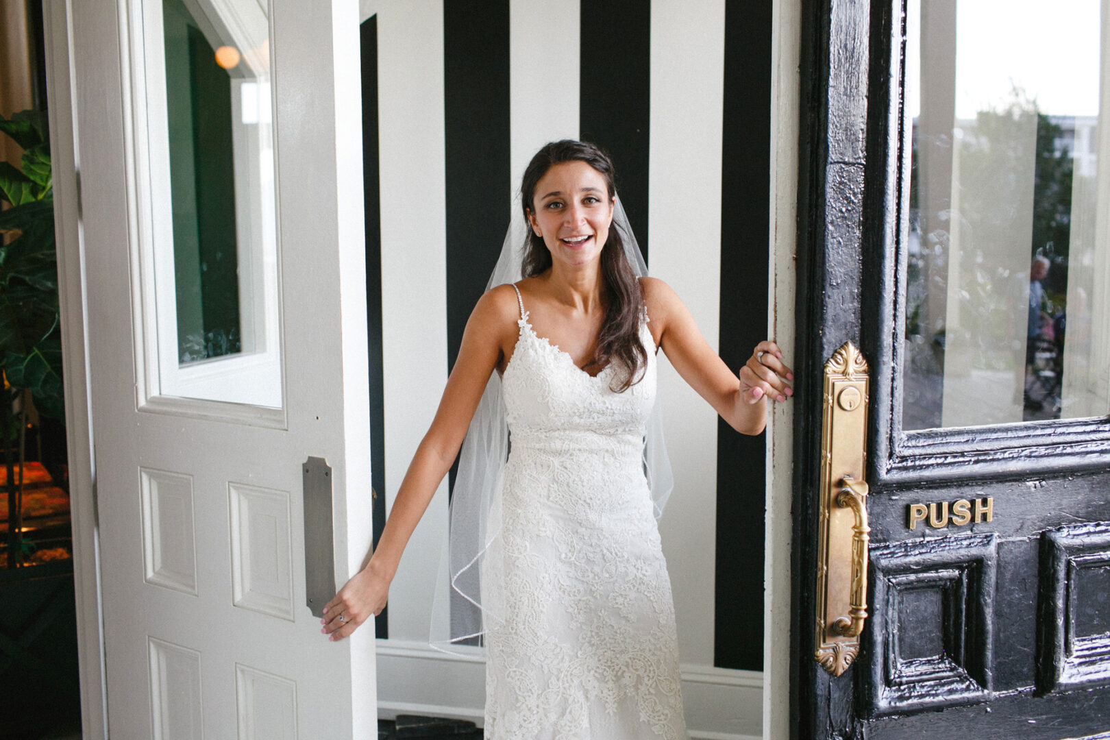 A bride in a white dress and veil smiles as she holds open a door with a "push" sign, standing gracefully against the backdrop of a striped wall at her charming Cape May wedding.