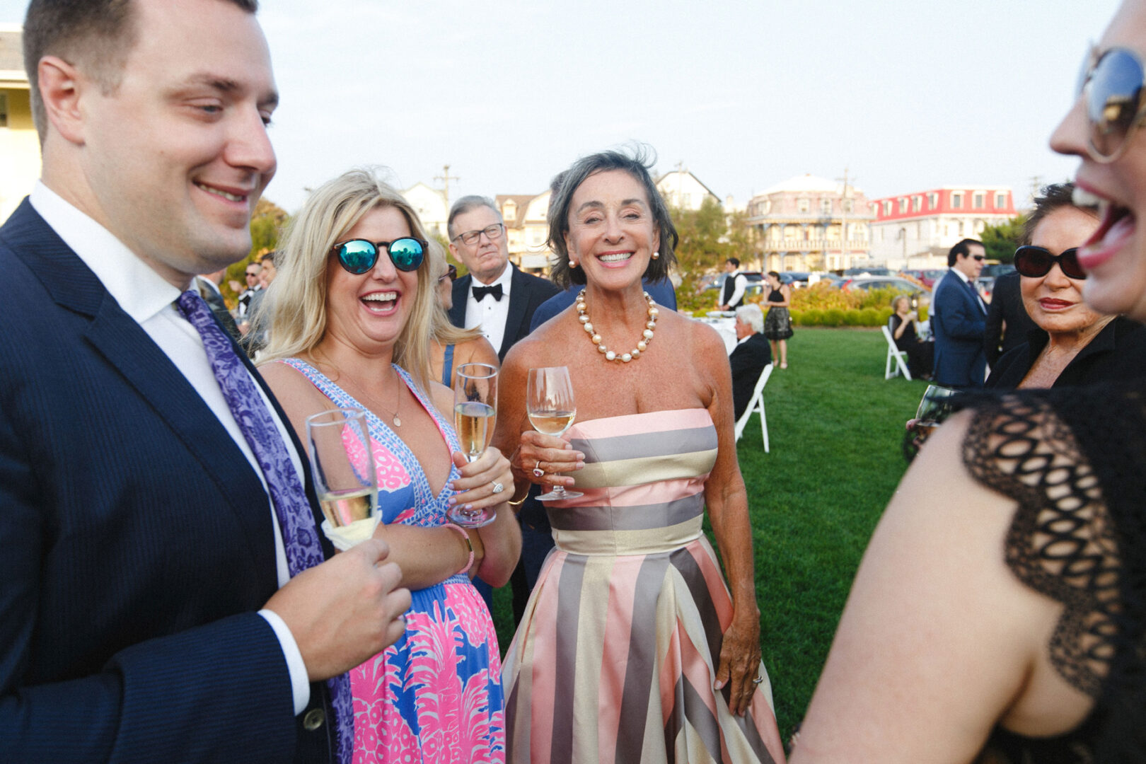 A group of people in formal attire are standing on a lush lawn in Cape May, holding champagne glasses and smiling, capturing the essence of a joyous wedding celebration.