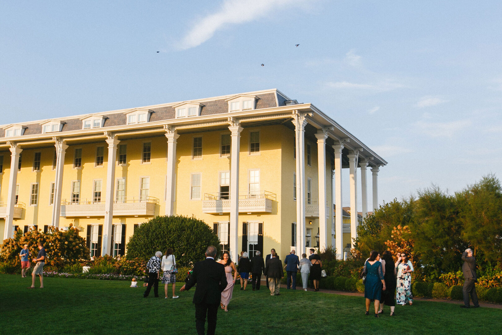 At a Cape May wedding, people dressed in formal attire gather on the lawn near a large, yellow, columned building under a clear sky.