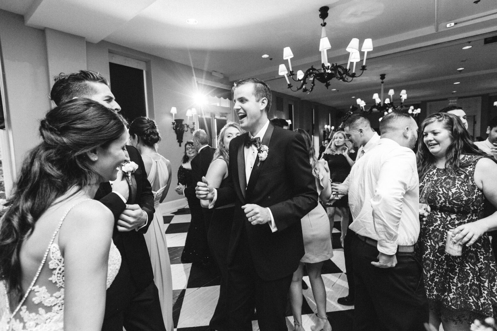 A black and white photo captures a Cape May wedding where elegantly dressed guests are laughing and socializing under dazzling chandeliers.