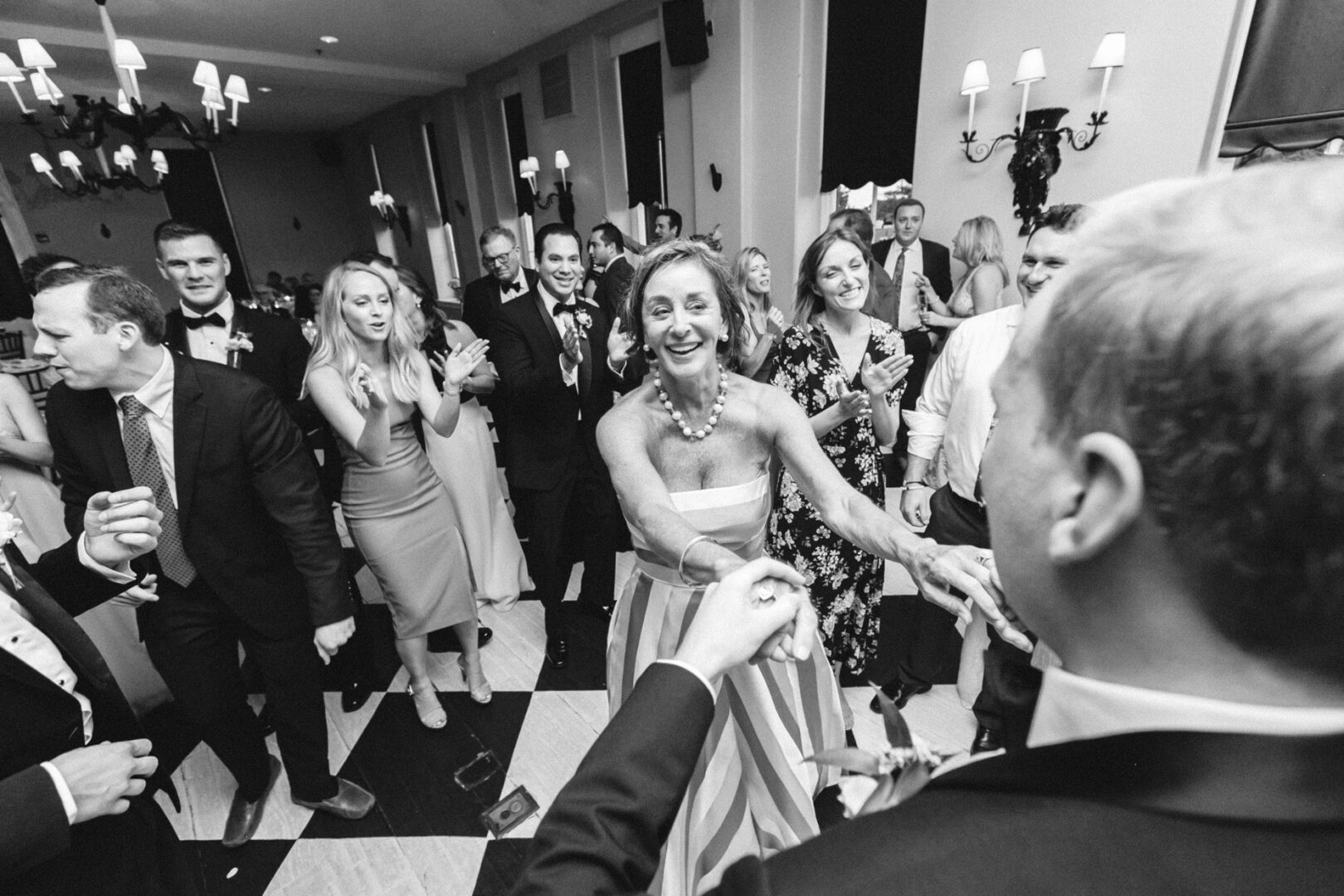 People dancing in a lively Cape May wedding scene, wearing formal attire. A woman in a striped dress dances with a man in the foreground. Black and white photo on a checkered floor.
