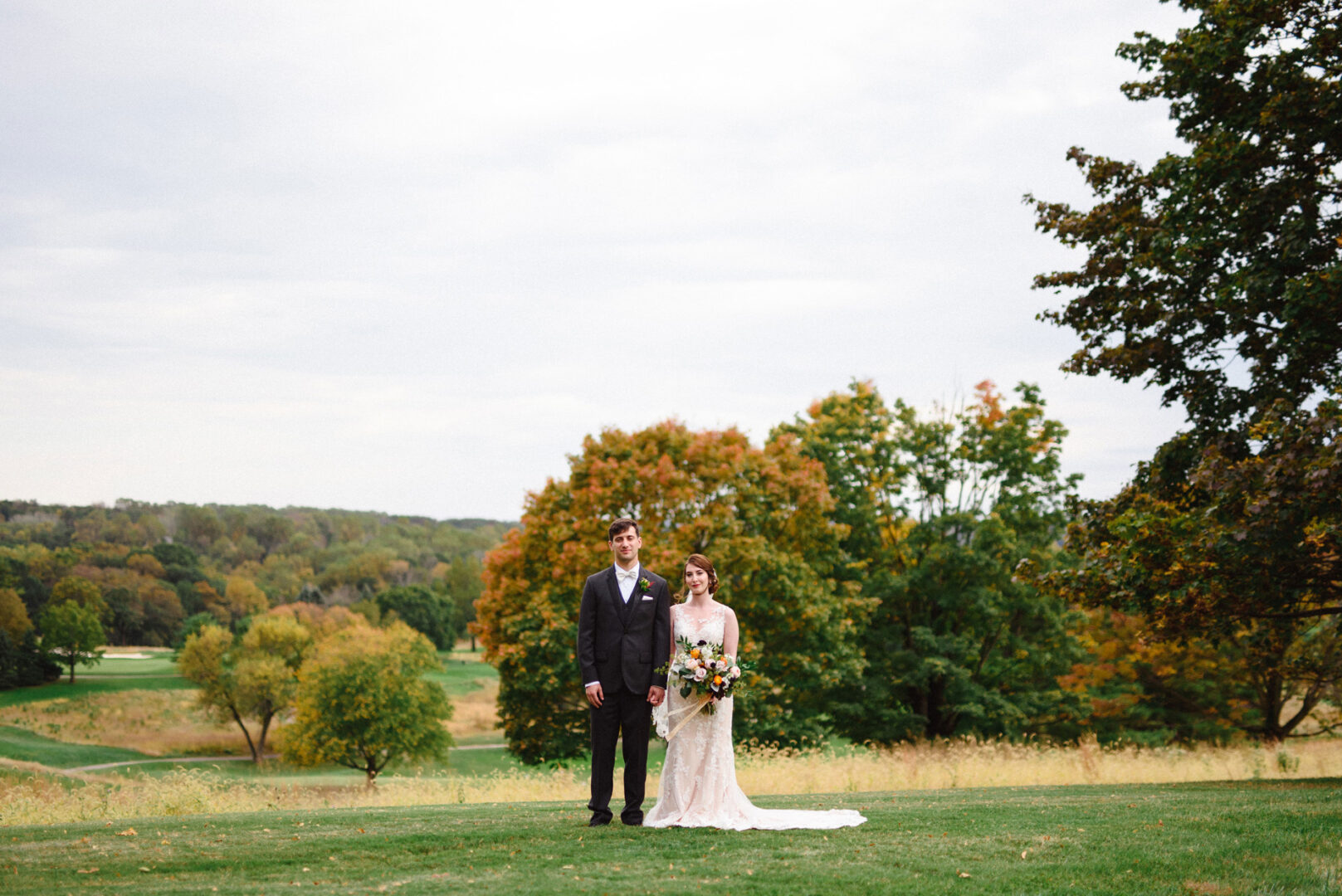 A couple in wedding attire stands on a grassy field at Union League at Liberty Hill, framed by trees and a cloudy sky in the background.