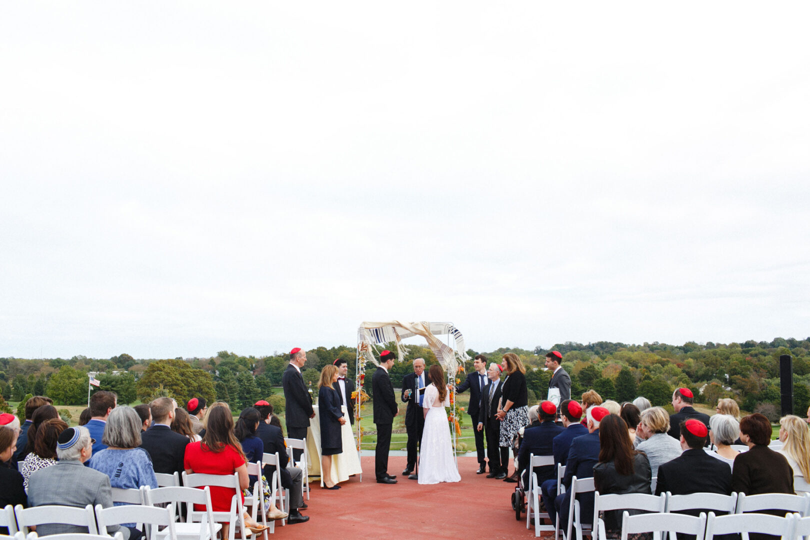 The Union League at Liberty Hill Wedding features an outdoor ceremony where guests sit on white chairs. Under a small canopy, the couple is surrounded by attendants and guests sporting red caps. Majestic trees and a cloudy sky provide a stunning backdrop to this elegant celebration.