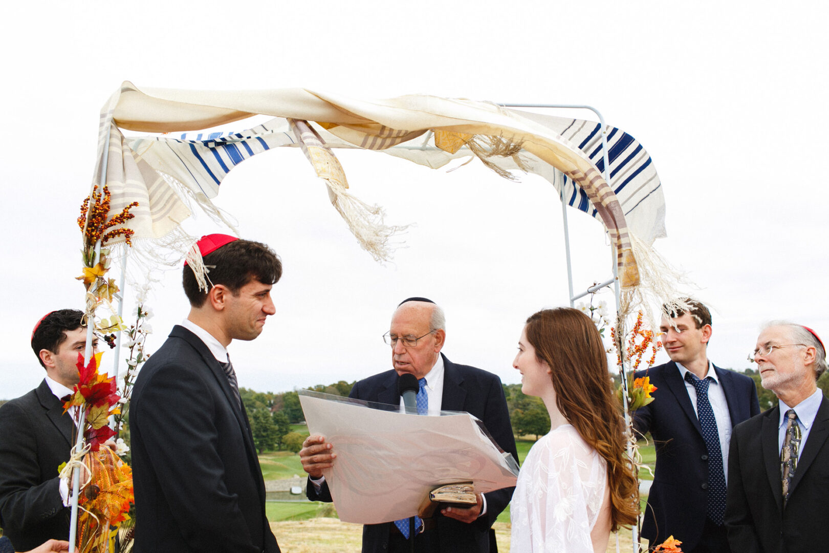 A group of people stand under a chuppah at the Union League at Liberty Hill, with a man reading from a document during a beautiful outdoor ceremony.