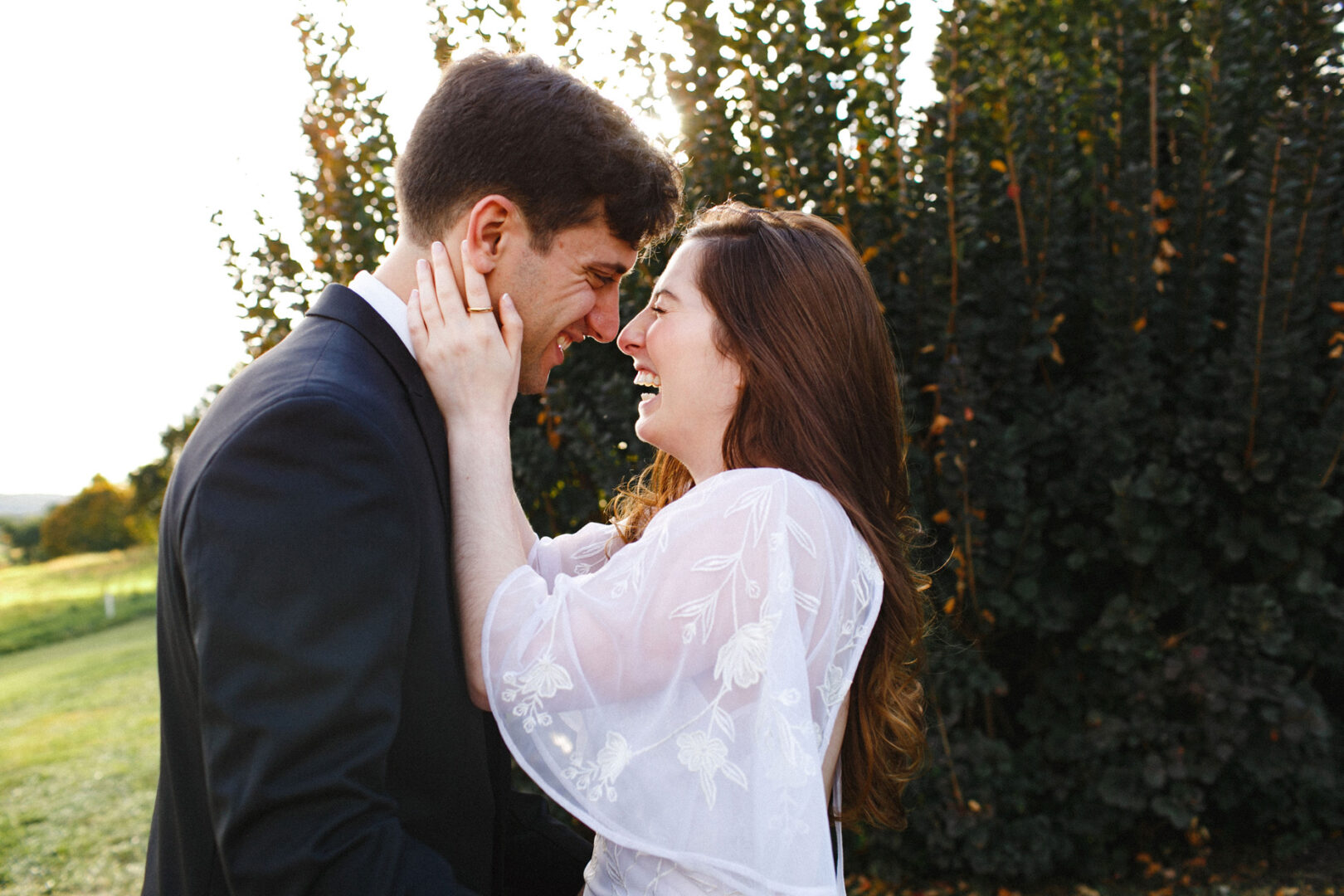 A couple stands close together outdoors at Union League at Liberty Hill, smiling at each other. The woman touches the man's face with both hands, surrounded by lush greenery.