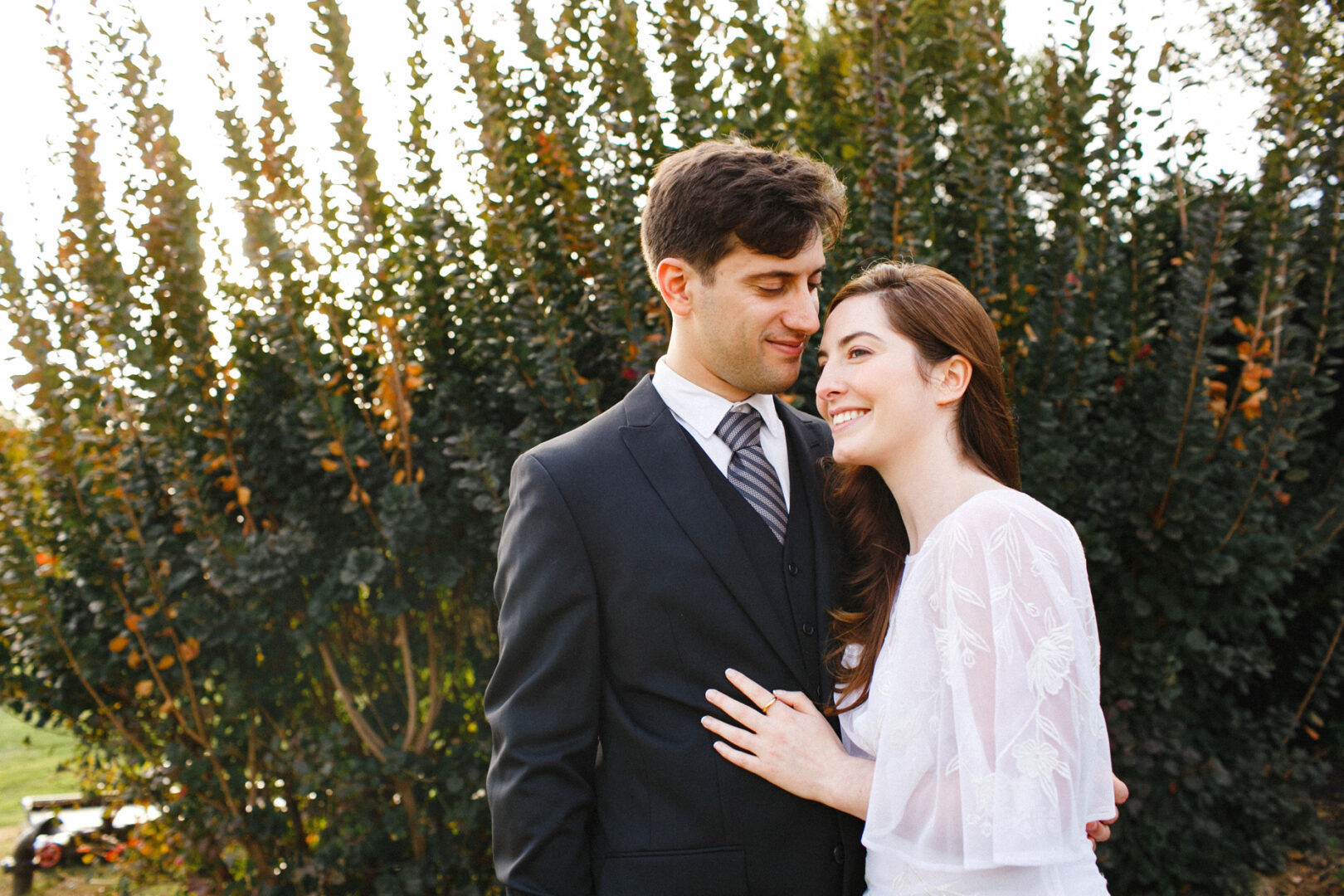 A couple stands together outdoors at their Union League at Liberty Hill wedding, smiling at each other. The man wears a suit and tie, and the woman is in a white dress. Lush greenery is in the background.