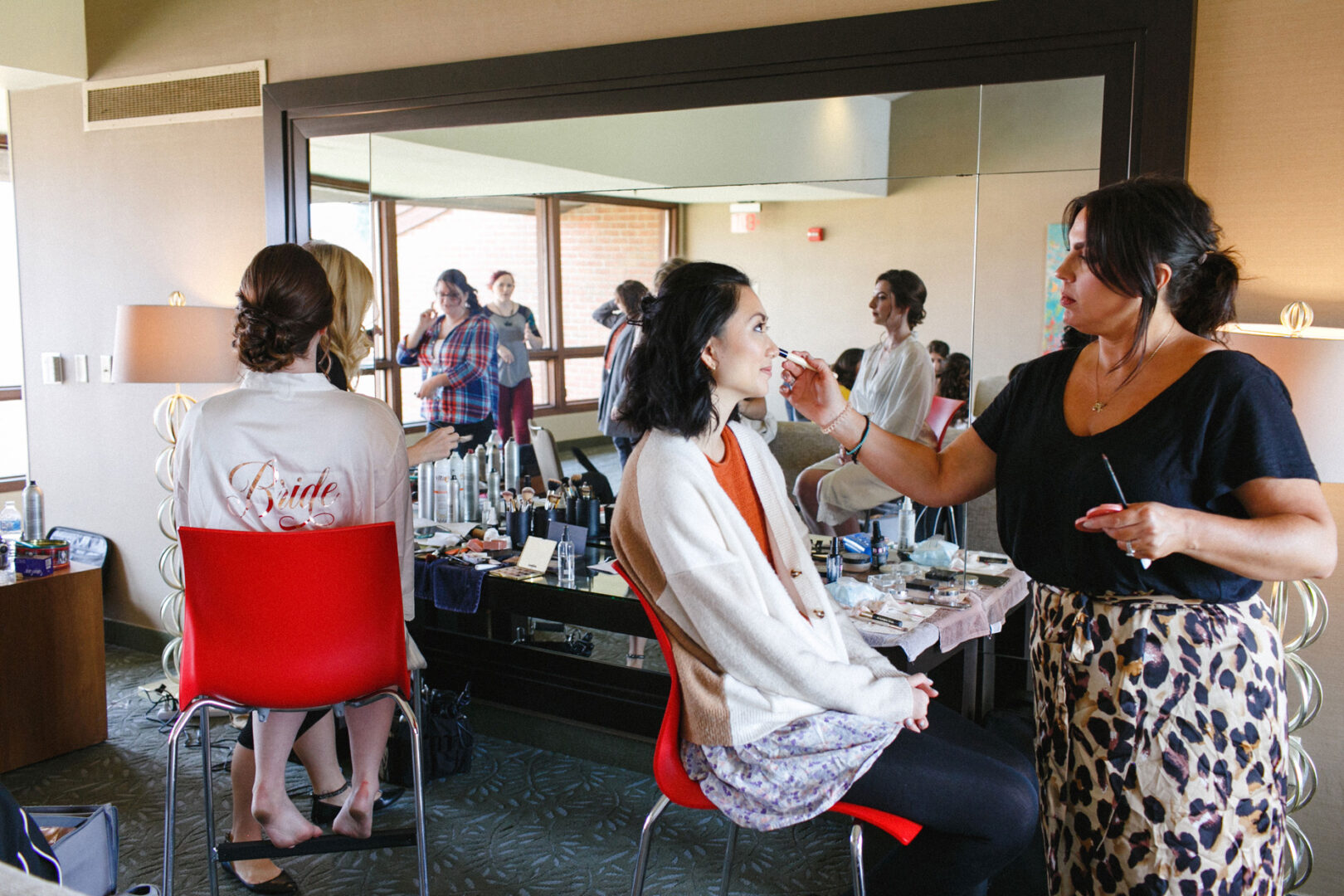 In a room with large mirrors, a woman carefully applies makeup to another seated woman. Nearby, a third woman, robed with "Bride" on her back, gazes out the window at the picturesque Union League at Liberty Hill wedding venue.