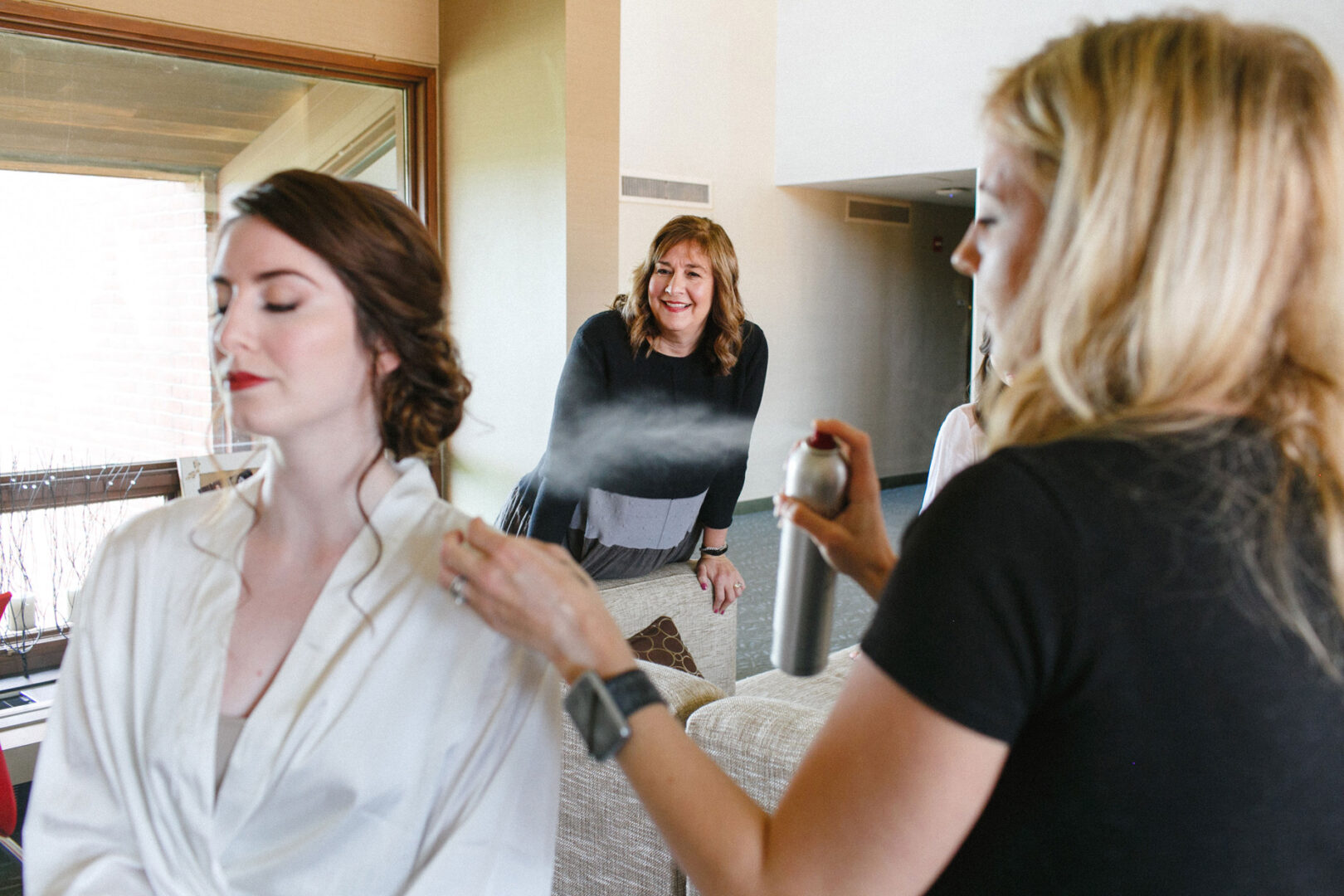 A woman in a white robe has her hair styled with hairspray by a hairstylist, preparing for the Union League at Liberty Hill wedding, while another woman smiles in the background.