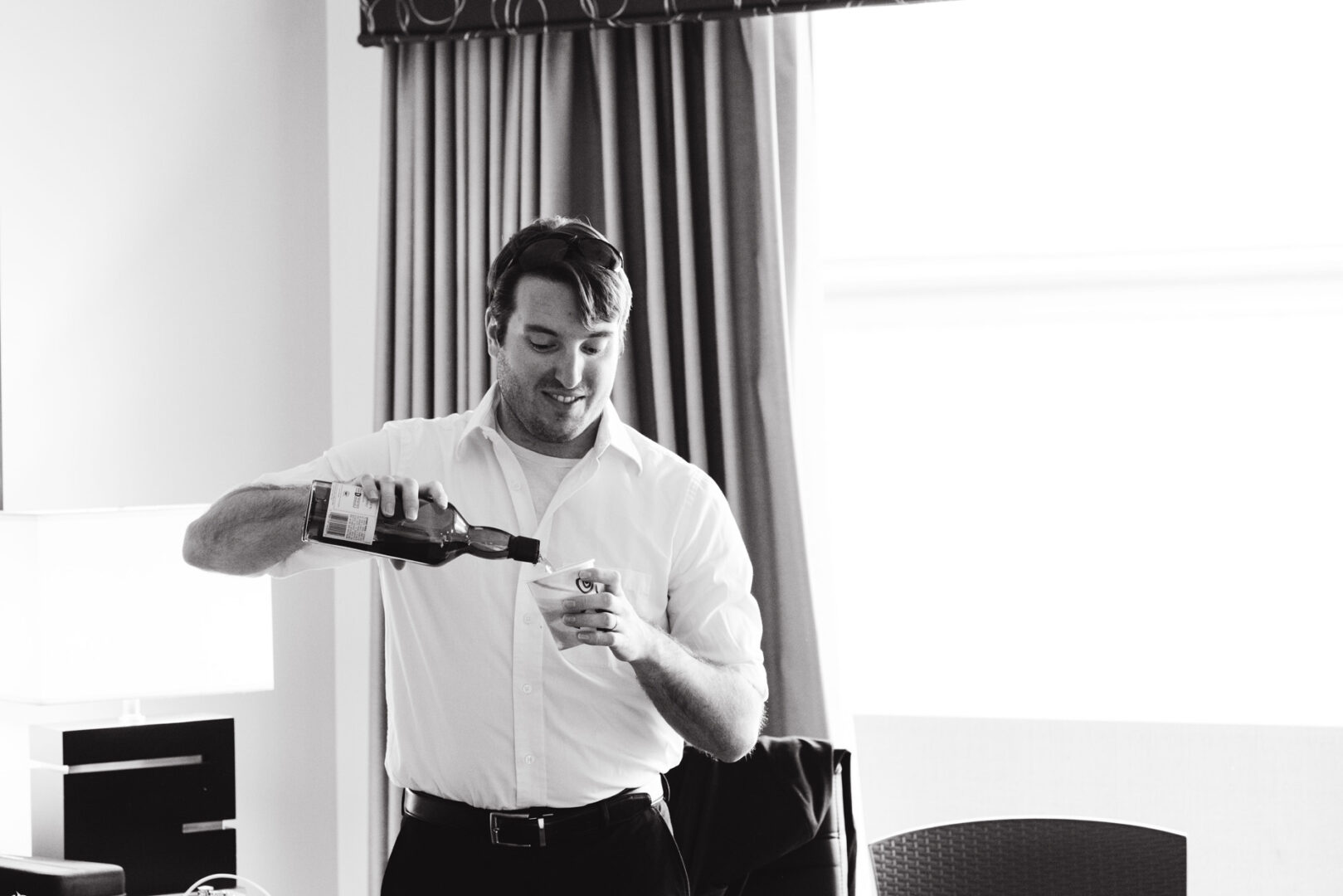 In a well-lit room with curtains and a lamp, a man in a white shirt pours whiskey into a cup, reminiscent of the elegant ambiance found at a Union League at Liberty Hill Wedding.