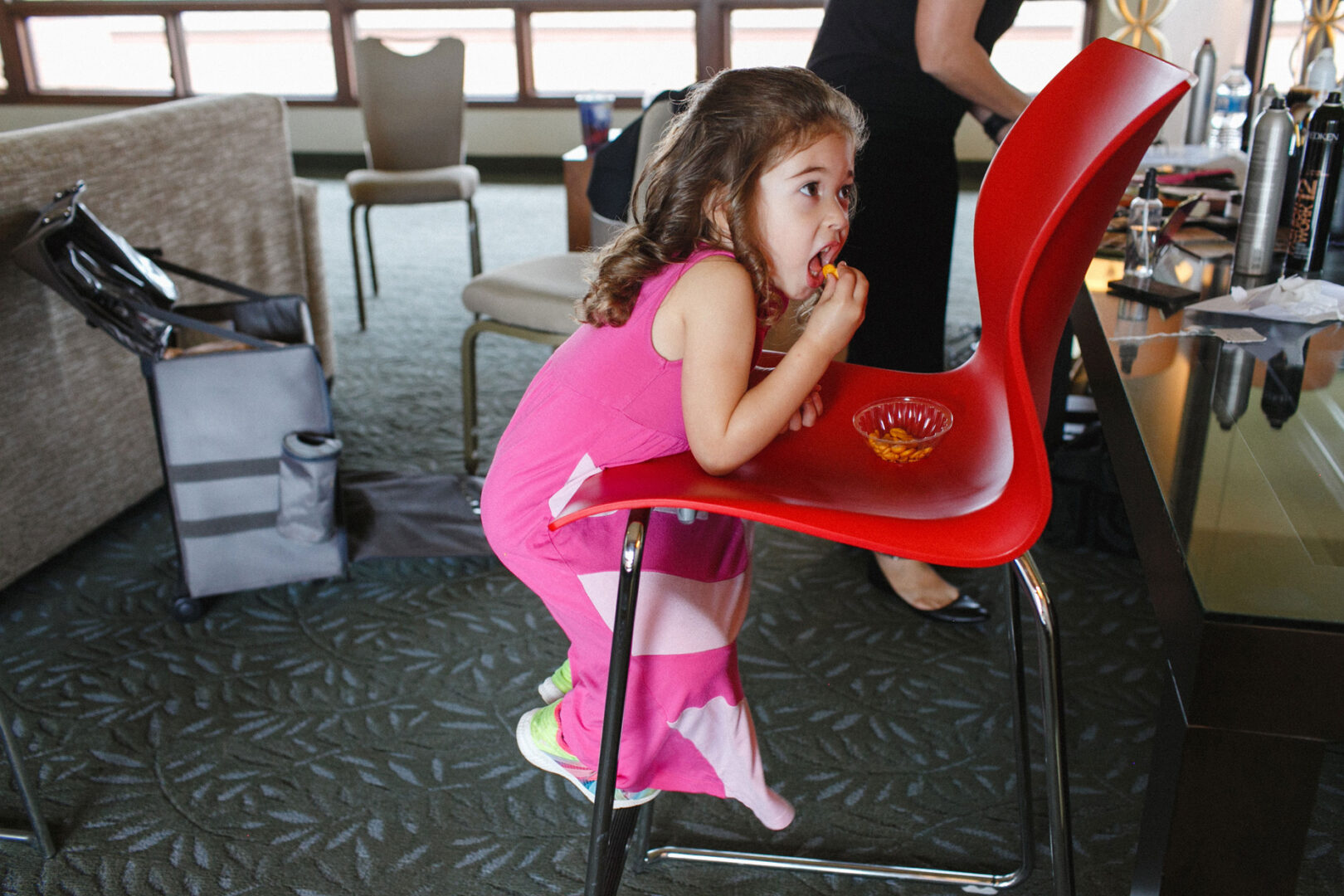 A young girl in a pink dress is leaning on a red chair, savoring bites from a small bowl. She stands indoors, as if amidst the elegant ambiance of a Union League at Liberty Hill Wedding, with various objects filling the background.