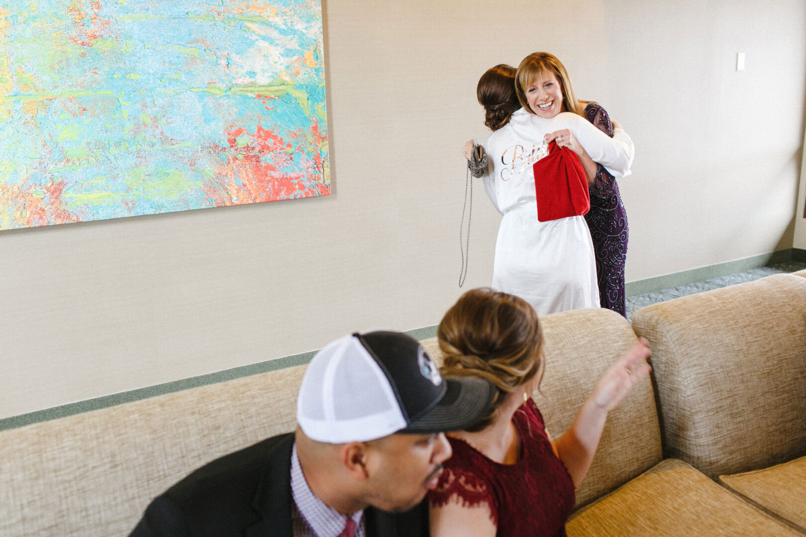 Two women embrace near a colorful painting, while a man in a baseball cap and a woman in a red dress sit on the couch in the foreground, capturing the joyful essence of a Union League at Liberty Hill Wedding.
