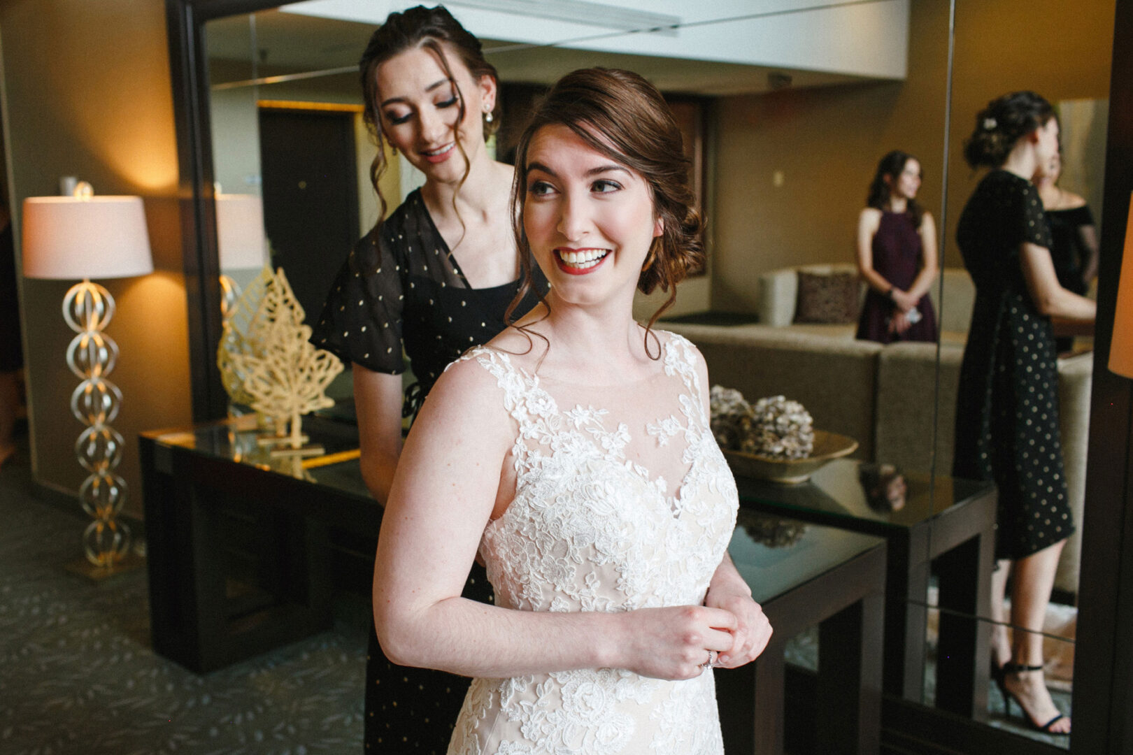 At a Union League at Liberty Hill Wedding, a bride in a lace dress smiles as a woman in black adjusts her outfit. Two others stand in the background of an elegantly decorated room with soft lighting from a vintage lamp.