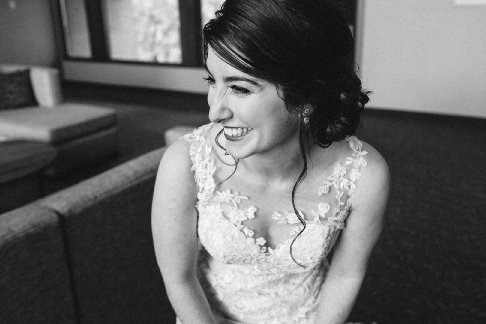 A woman in a lace wedding dress sits on a sofa at the Union League at Liberty Hill, smiling and gazing to the side in a timeless black and white photo.