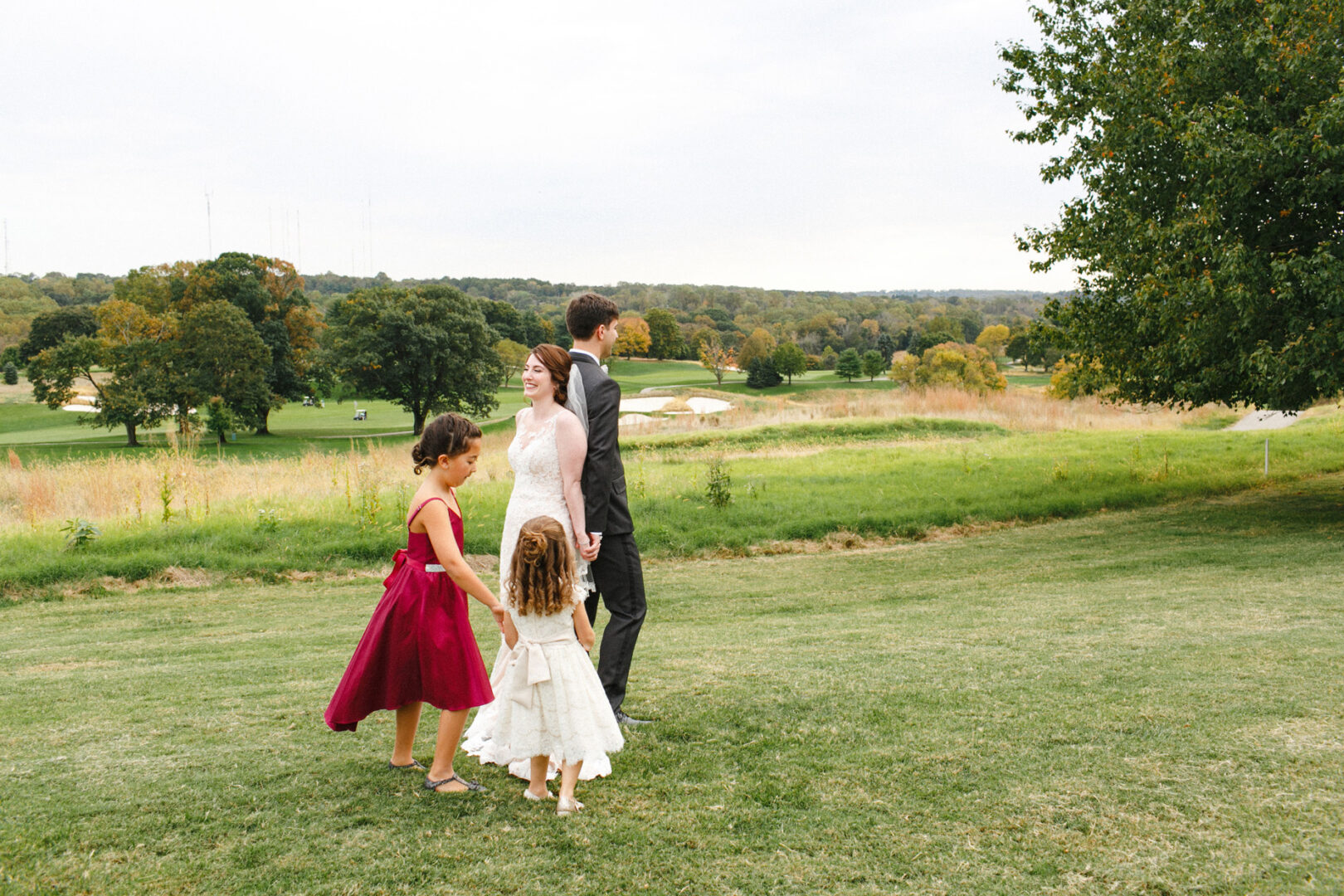 A bride and groom hold hands with two young girls in formal dresses, strolling across the grassy fields of the Union League at Liberty Hill, with trees gracefully lining their path.