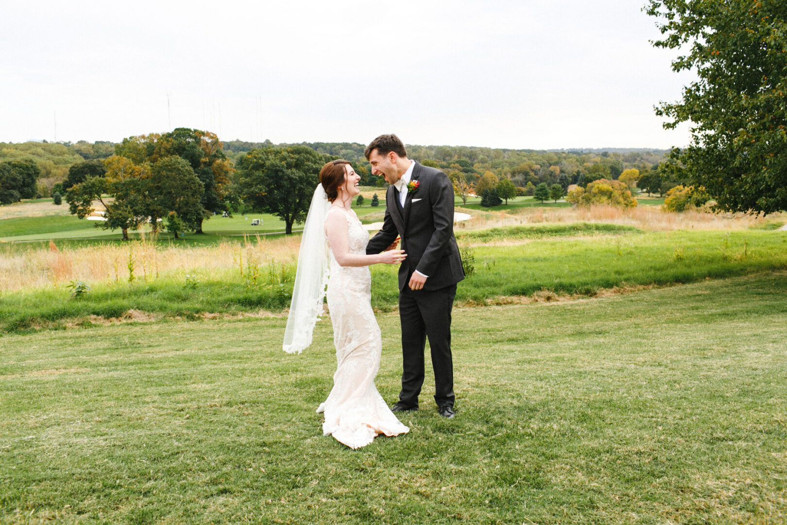 A bride and groom exchange warm smiles on a grassy field at Union League Liberty Hill, surrounded by verdant trees and rolling hills in the background.