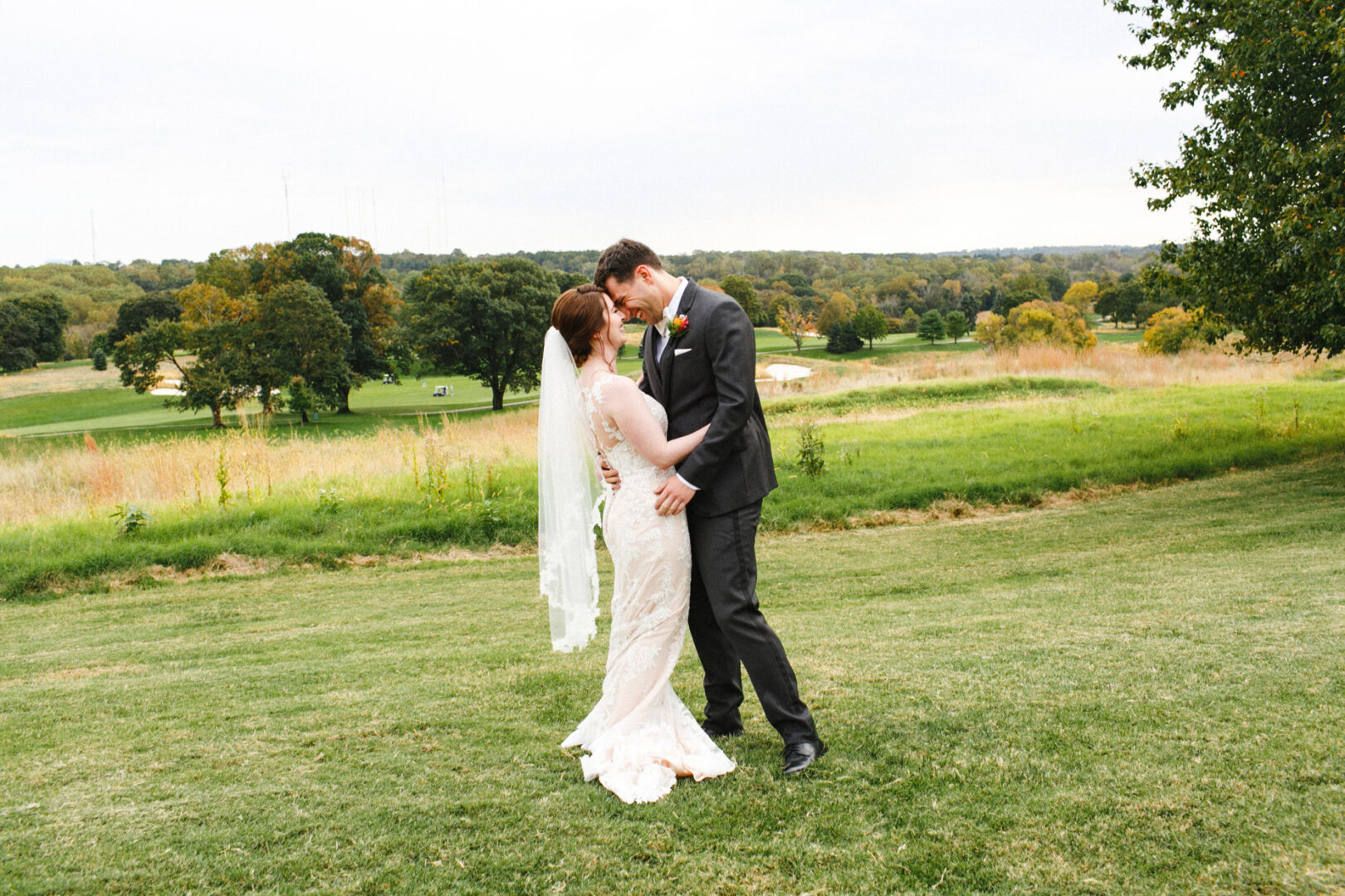 At their Union League at Liberty Hill wedding, a bride and groom share a kiss on a grassy field, surrounded by trees and a rolling landscape in the background.