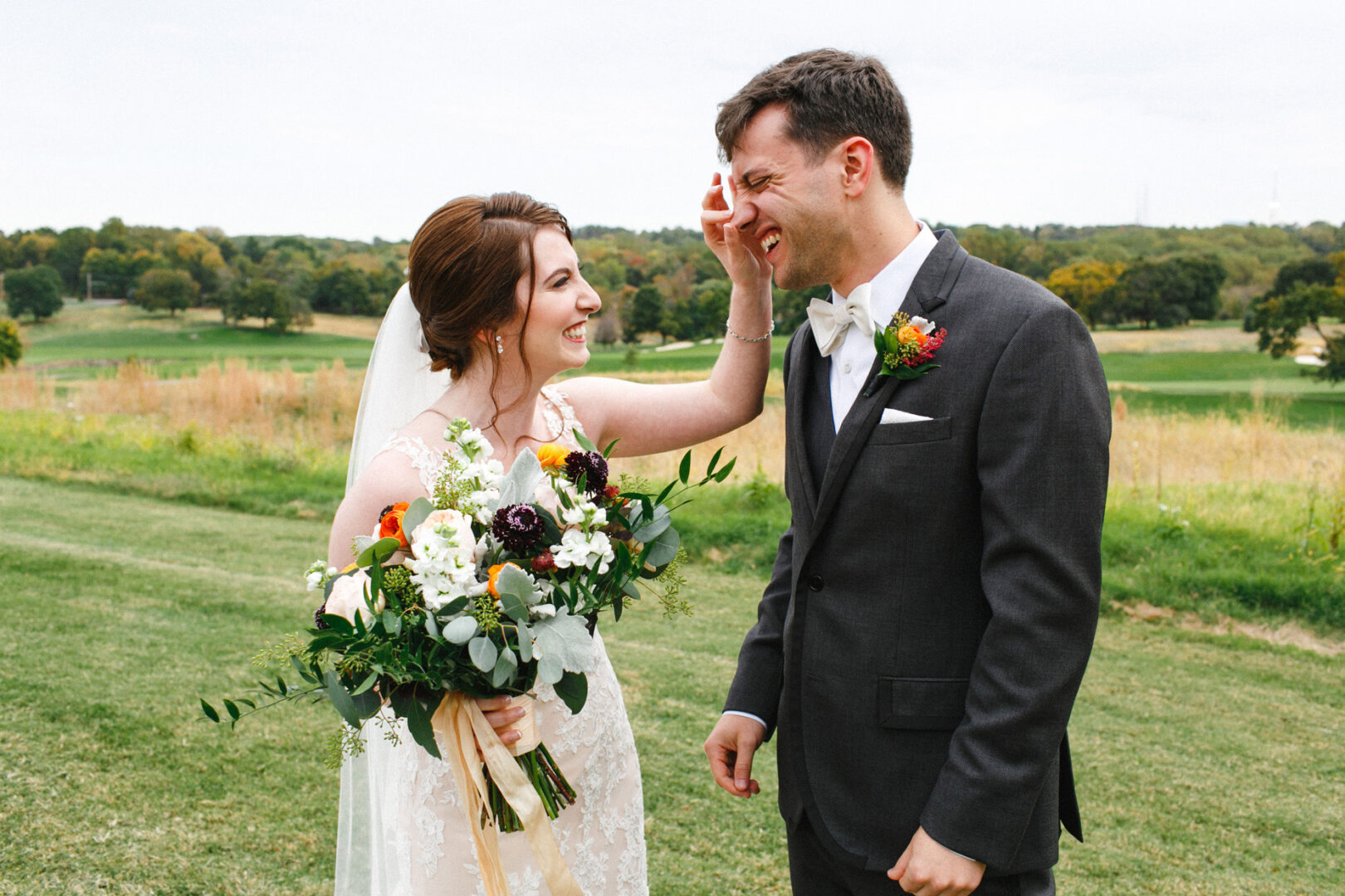 A bride and groom stand on a grassy field at the Union League at Liberty Hill Wedding. The bride, holding a bouquet, gently touches the groom's face as they both smile, surrounded by lush trees and grass in the background.