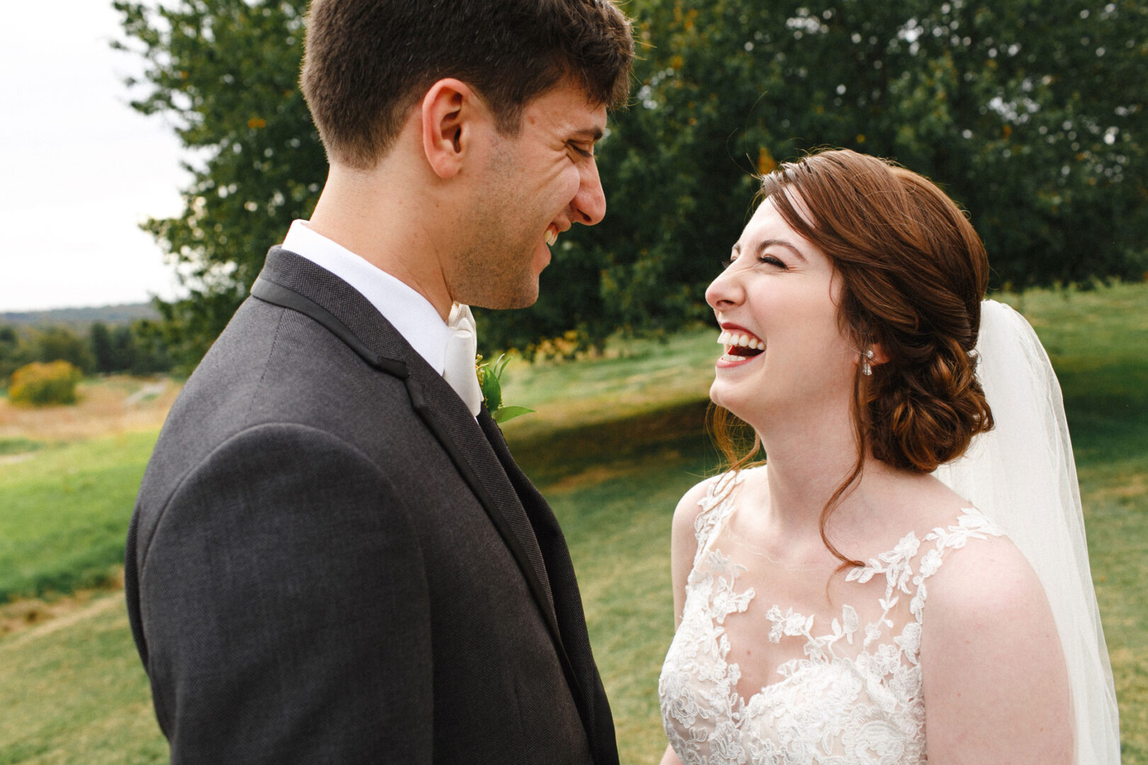 A bride and groom smile at each other outdoors amidst the lush greenery of their Union League at Liberty Hill wedding. The bride wears a lace dress and veil, while the groom is in a dark suit, perfectly capturing their enchanting moment.