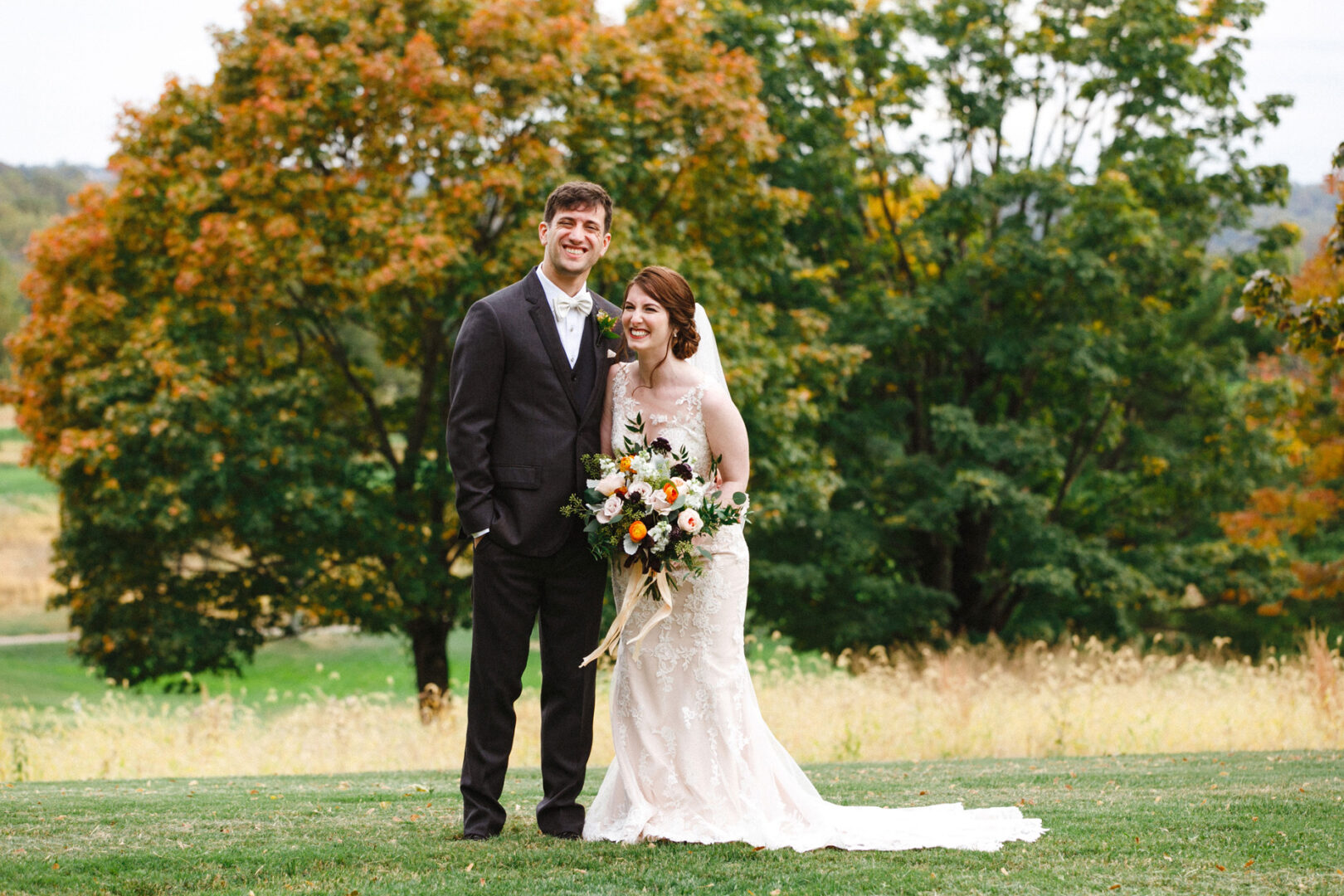 A bride in a white gown holds a bouquet beside a groom in a dark suit at their Union League at Liberty Hill wedding. They smile on a grassy area with trees showing fall foliage in the background.