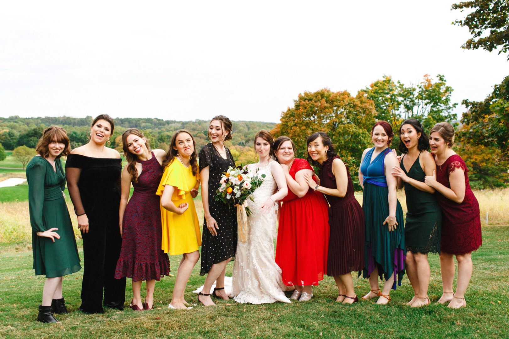 A bride in a white dress poses outdoors at the Union League at Liberty Hill Wedding with eleven bridesmaids in vibrant dresses, standing in a row on a grassy area framed by trees.