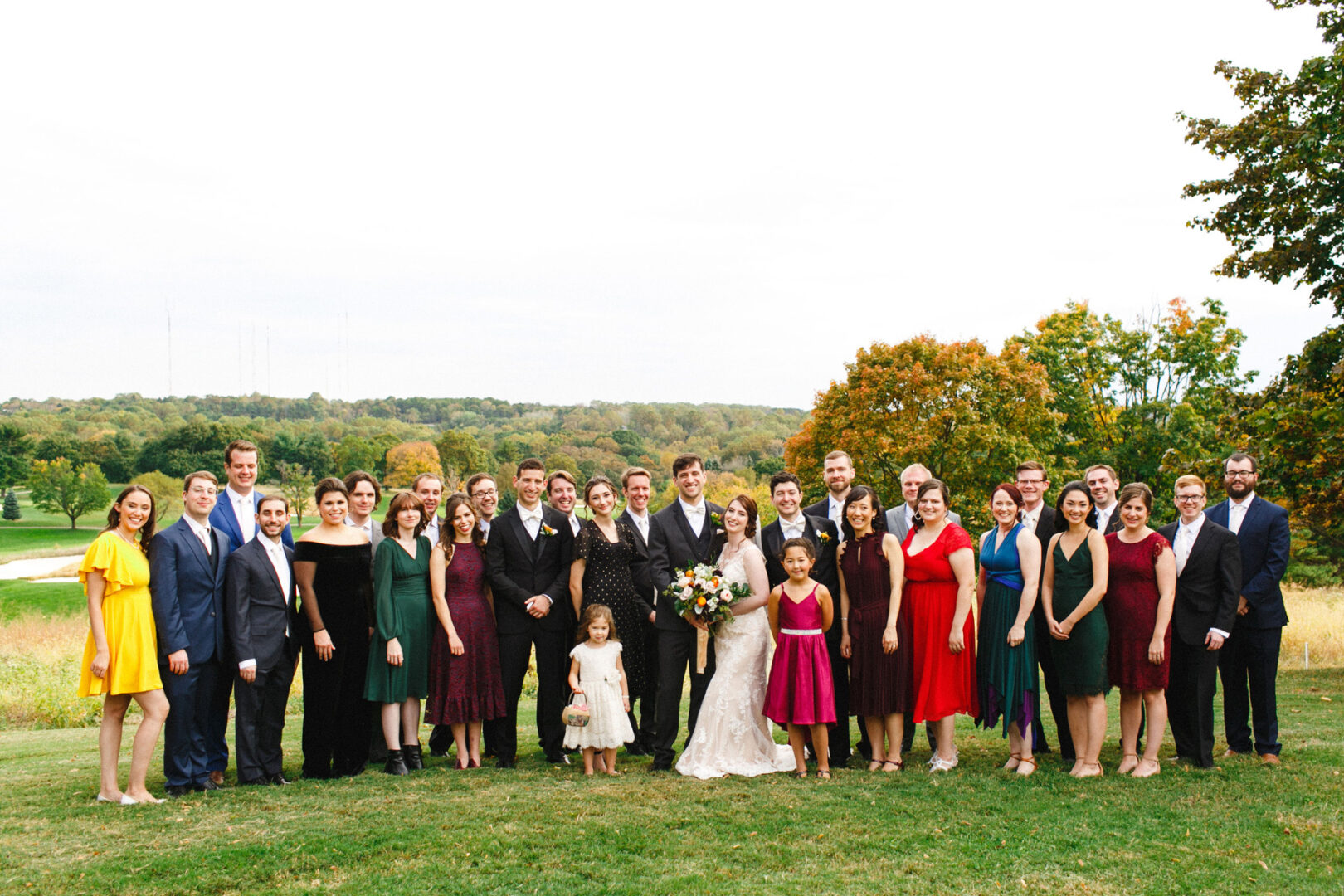 A large group of people stands on the grass in formal attire, centered around a bride and groom at the scenic Union League at Liberty Hill wedding. Trees and a picturesque landscape provide a stunning backdrop to this elegant celebration.