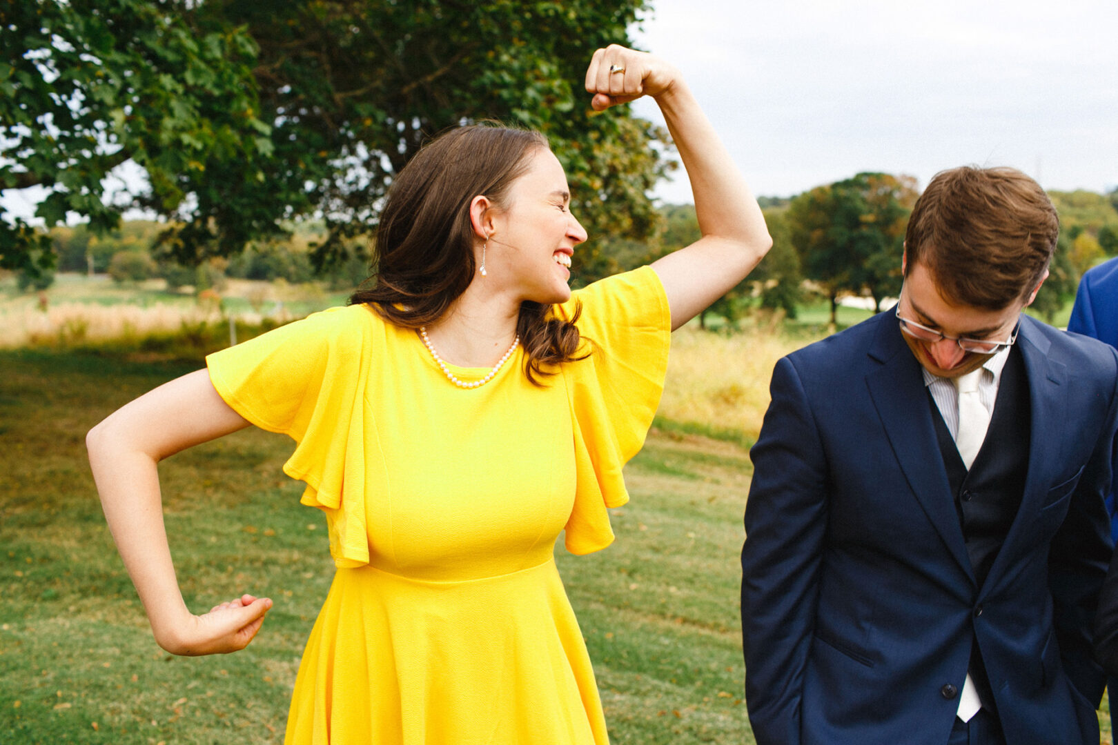 At a Union League at Liberty Hill wedding, a woman in a yellow dress playfully flexes her arm alongside a smiling man in a suit, savoring the joyous outdoor celebration.