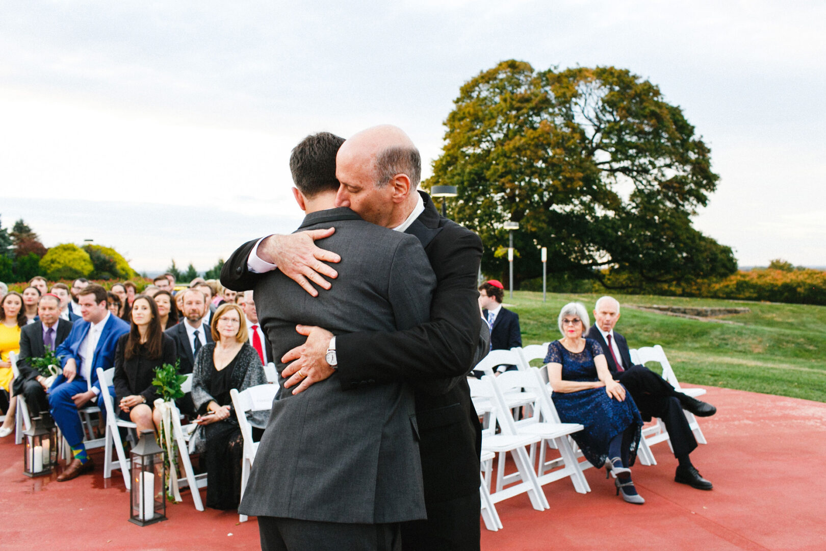 Two men in suits embrace at an outdoor wedding event, held at the picturesque Union League at Liberty Hill, with seated attendees, a lush green lawn, and a majestic large tree providing a serene backdrop.