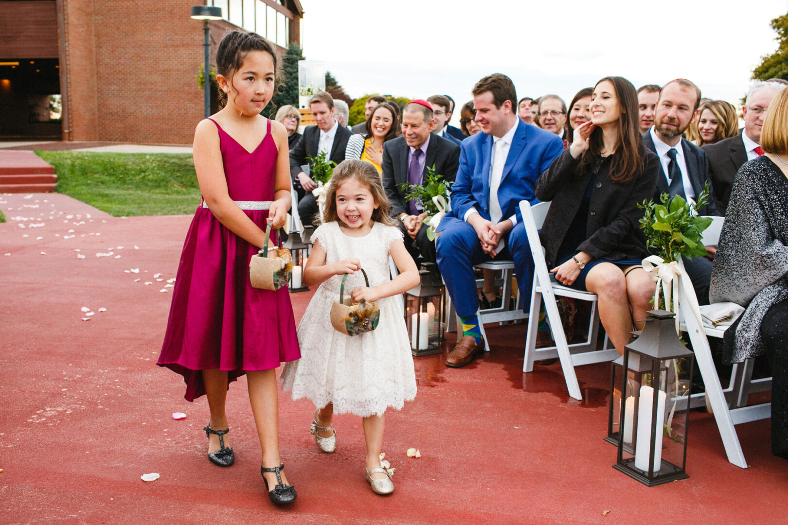 Two young girls, one in a red dress and the other in white, gracefully walk down the outdoor aisle at a Union League at Liberty Hill wedding, carrying baskets. Seated guests, dressed in formal attire, watch attentively.
