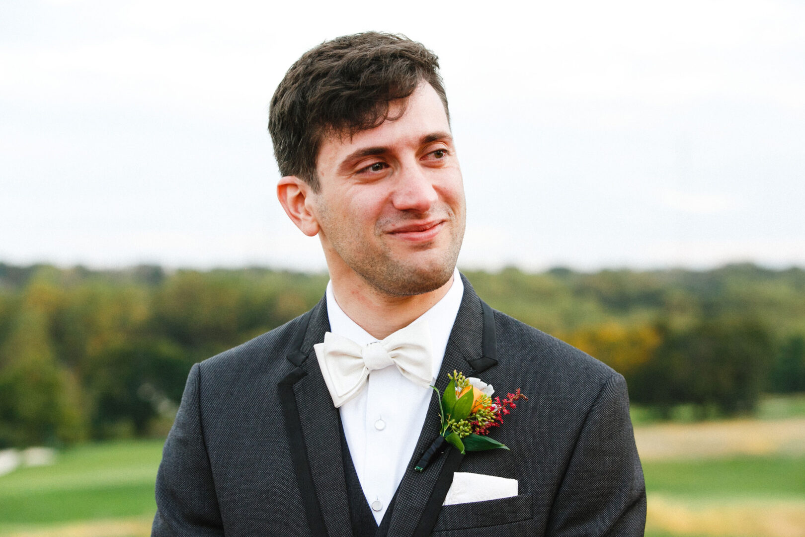 A man in a tuxedo with a white bow tie and boutonniere smiles slightly while standing outdoors at the Union League at Liberty Hill. Trees and grass are in the blurred background, capturing the serene charm of a wedding setting.