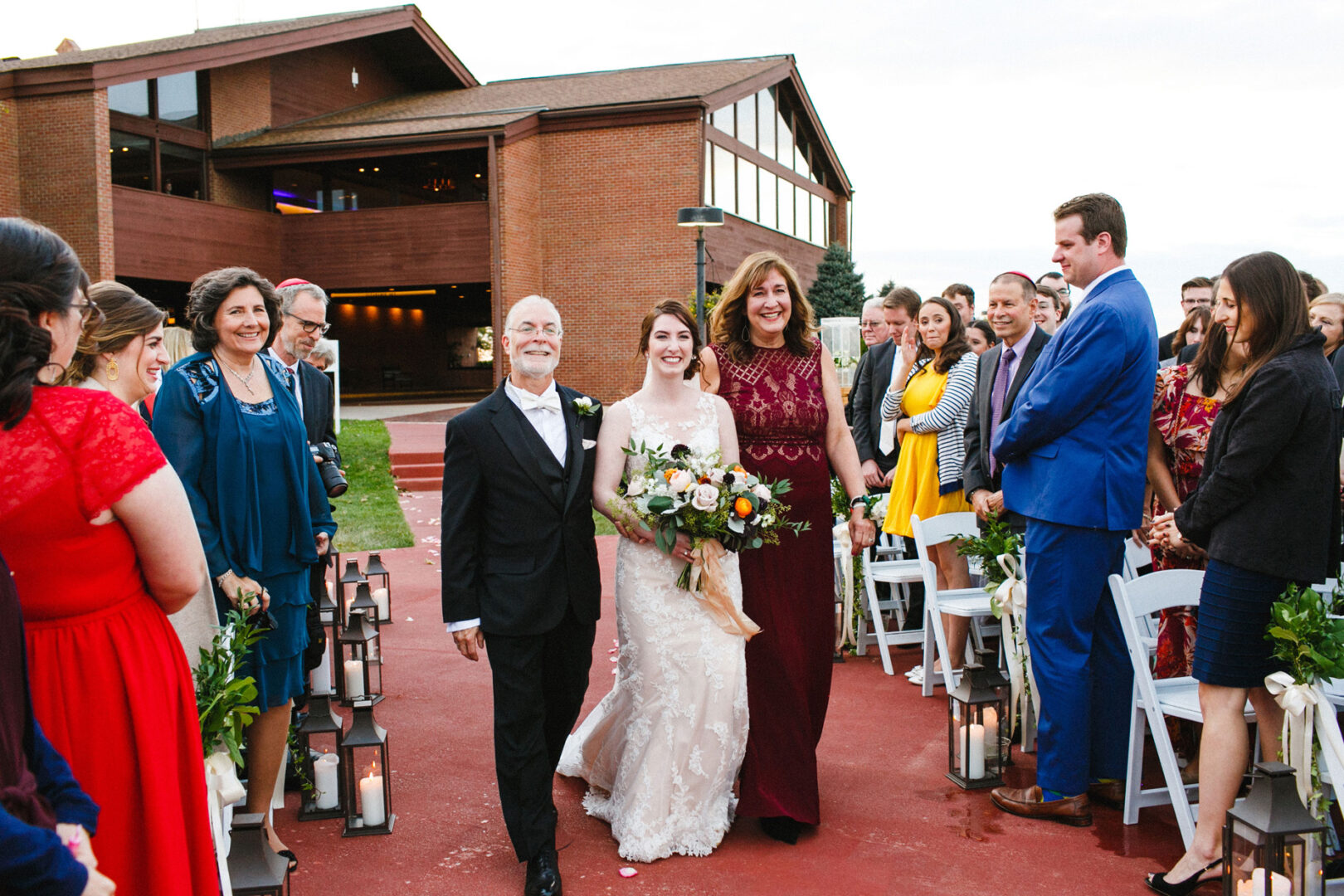 At a stunning Union League at Liberty Hill wedding, a bride in a white dress gracefully walks down the aisle outdoors, flanked by two escorts. Guests are seated on either side, with the elegant brick building serving as a picturesque backdrop to this unforgettable moment.