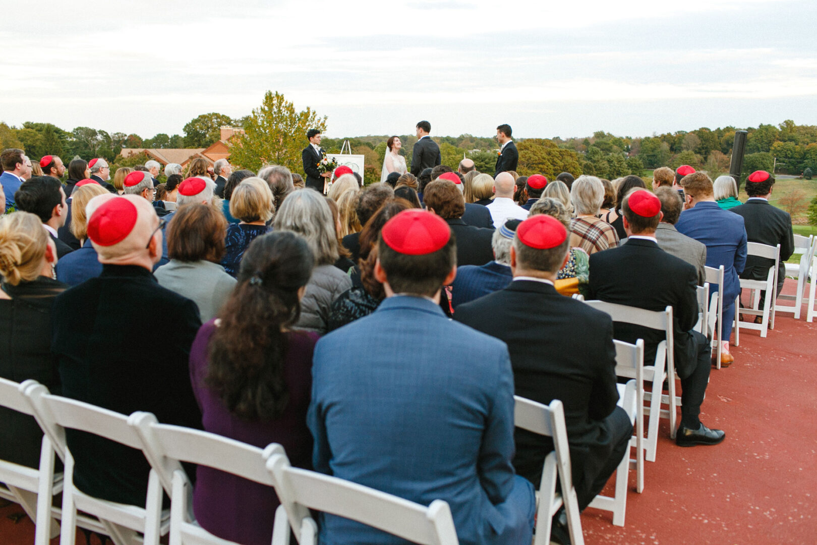 At the Union League at Liberty Hill, an outdoor wedding ceremony unfolds with attendees in red head coverings seated on white chairs. The bride and groom stand gracefully at the altar, embraced by towering trees and beneath a vast open sky.