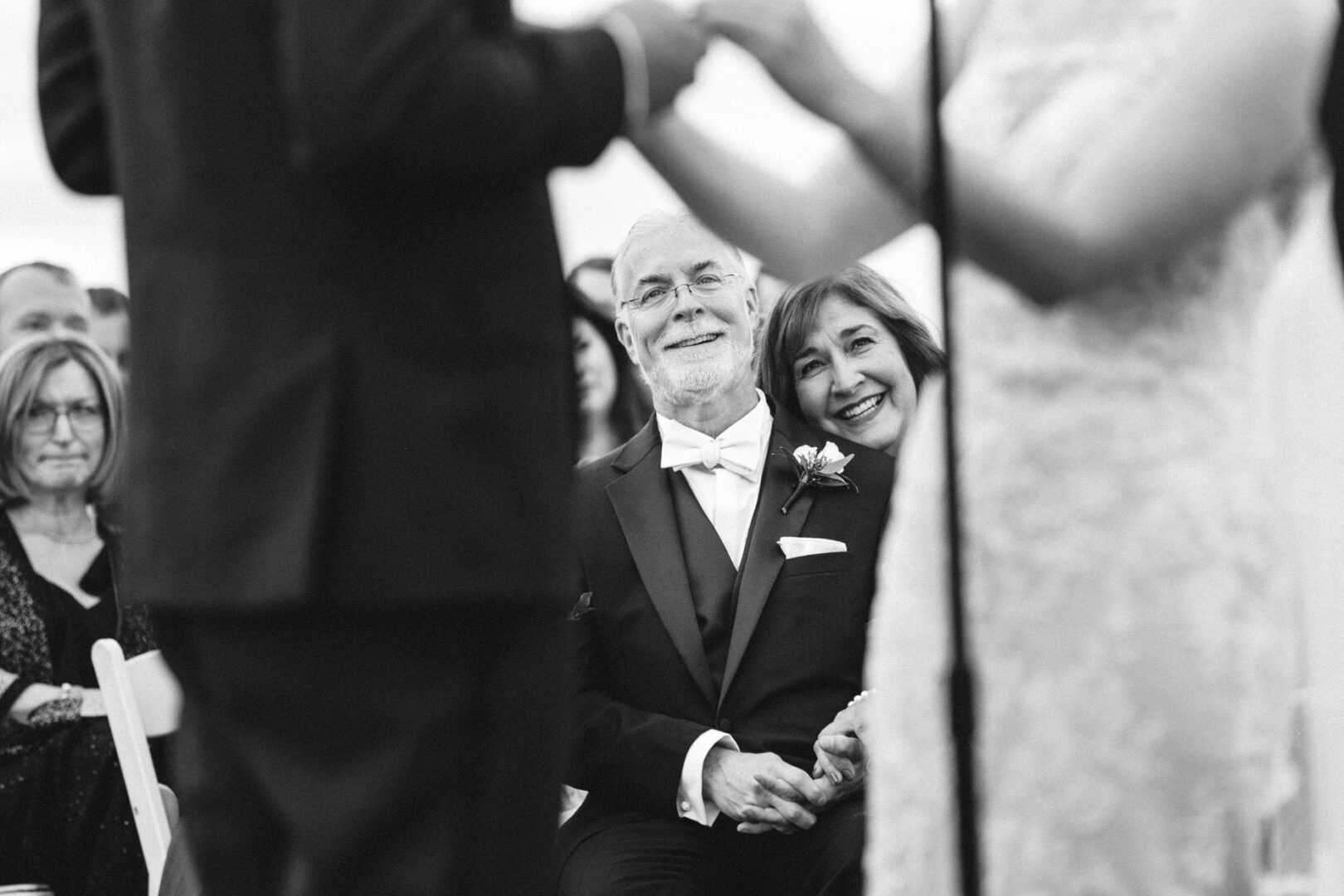 A couple sits smiling and holding hands, watching a Union League at Liberty Hill wedding ceremony. The man in a suit and the woman leaning on him create an intimate moment, captured beautifully in black and white.