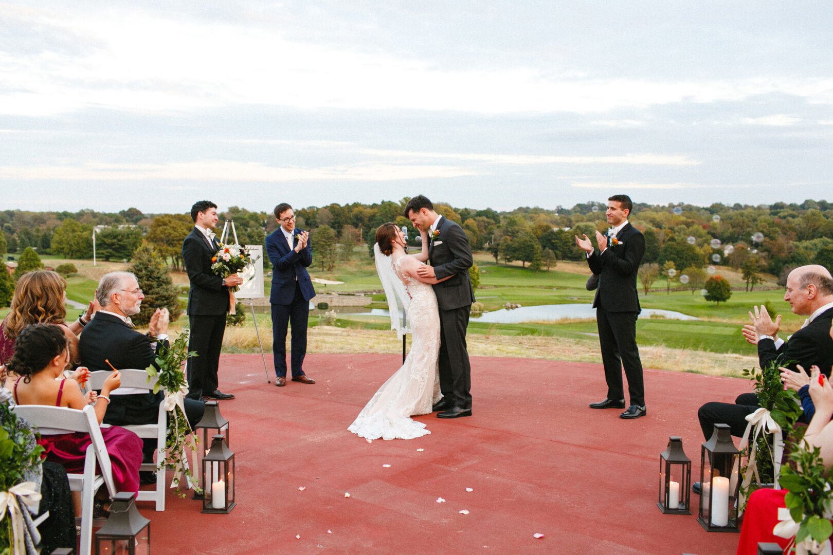 A couple kisses during their Union League at Liberty Hill wedding ceremony, surrounded by the bridal party and seated guests, with a scenic landscape in the background.