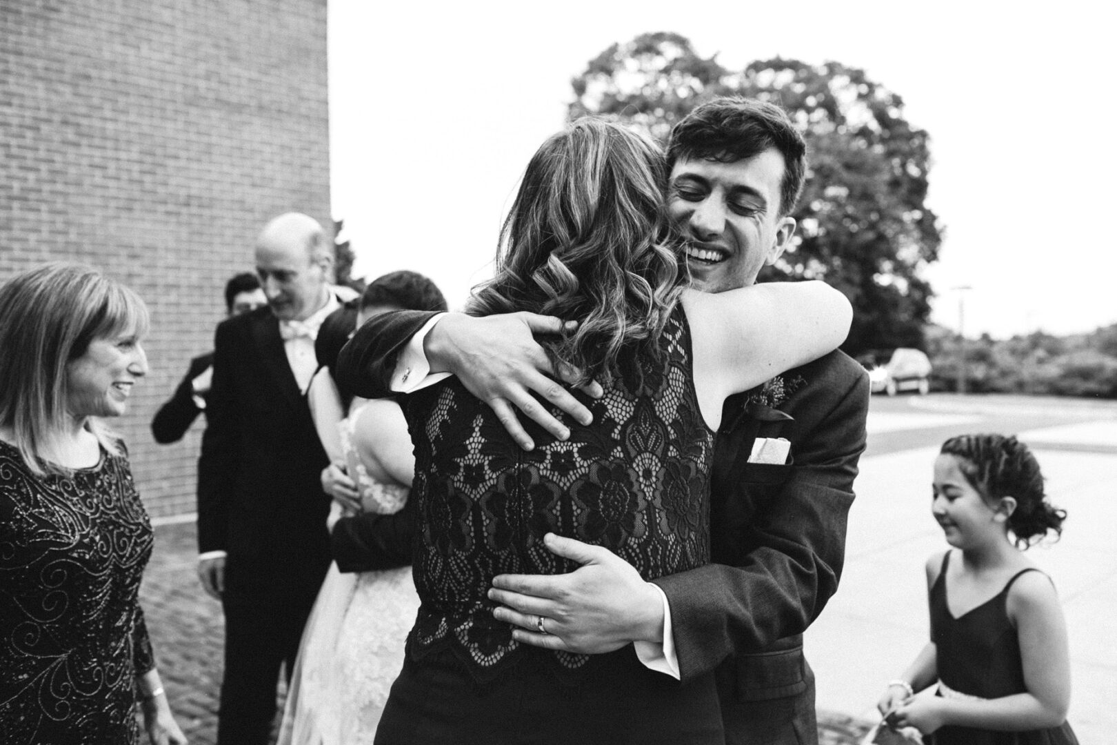 A man in a suit and a woman in a lace dress share an embrace outdoors at the Union League at Liberty Hill wedding. Nearby, others, including a girl, are engaged in lively conversations.