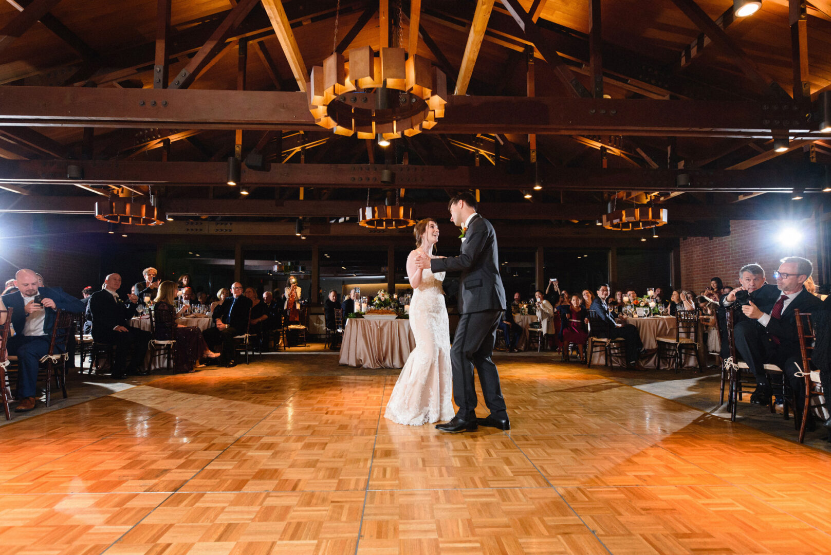 At the Union League at Liberty Hill, a couple dances gracefully in a warmly lit venue, surrounded by guests seated around the elegant wooden dance floor.