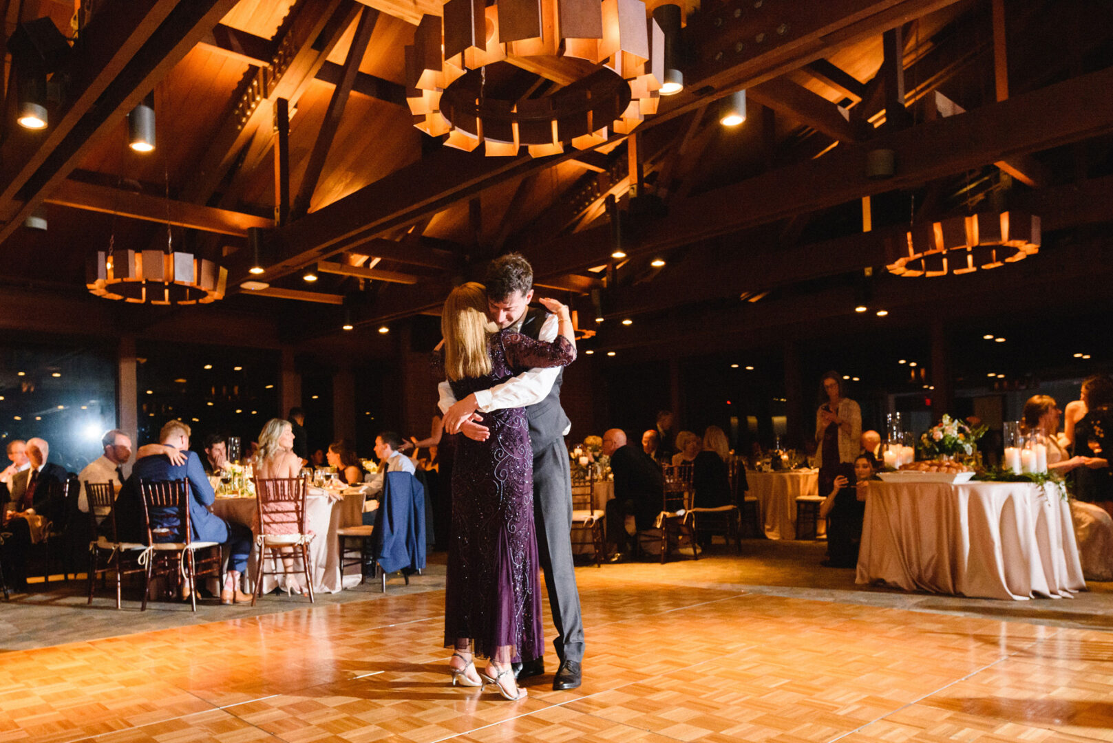 At the Union League at Liberty Hill, a couple dances closely on a wooden floor in an elegant event hall. Guests are seated at round tables around them, while warm lighting creates a cozy ambiance from above.