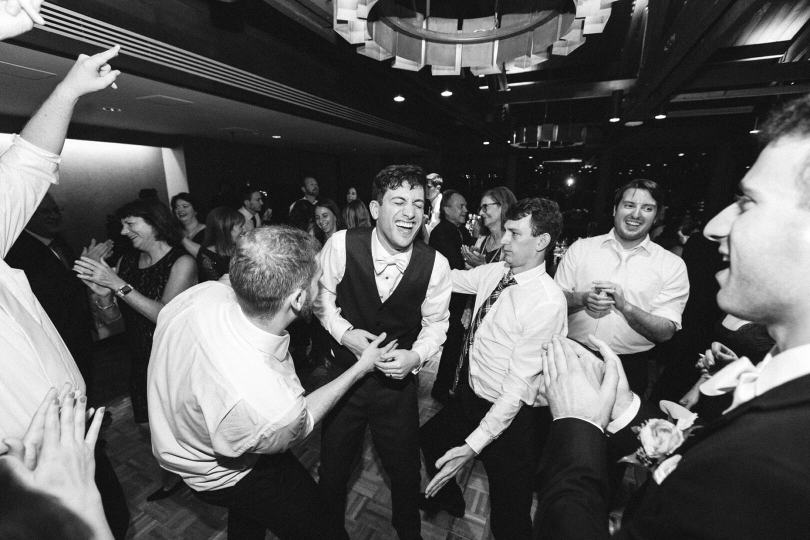 A joyful group is dancing and laughing at a lively indoor event, with one person at the center wearing a suit and bow tie. The scene captures the elegance of a Union League at Liberty Hill wedding, where celebration sparks connections and unforgettable memories are made.