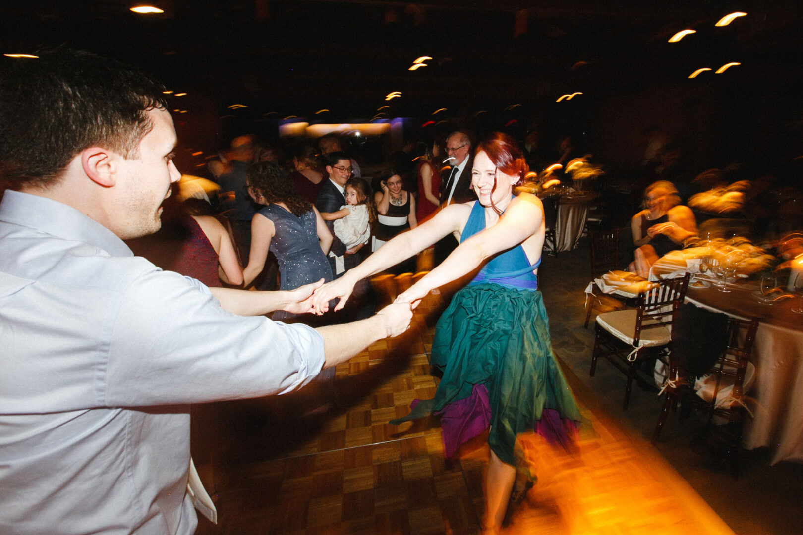Amidst the dimly lit ambience of the Union League at Liberty Hill Wedding, two people danced energetically, joined by other couples and seated guests in the background, celebrating love's joyful rhythm.