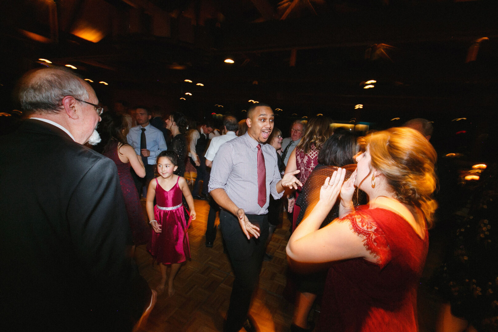 At a vibrant Union League at Liberty Hill Wedding, a group of people danced energetically indoors. Focus shifts to a man in a shirt and tie, expressing pure excitement, while others, including a young girl in a dress, are seen smiling and joyfully moving around him.