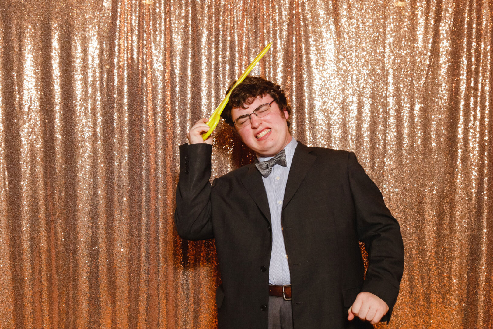 At a Union League at Liberty Hill wedding, a man in a suit poses playfully with a yellow inflatable sword against a sparkling gold backdrop.