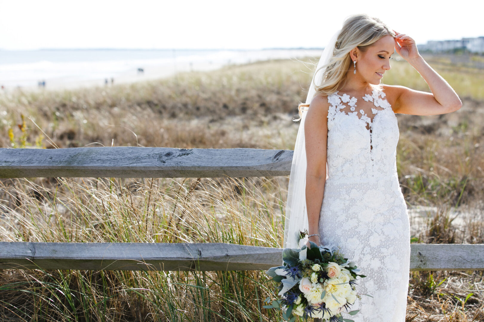 The bride, in a white lace dress, holds a bouquet while standing near a wooden fence on the grassy beach. The serene sea forms the backdrop to this enchanting Yacht Club of Sea Isle City wedding scene.