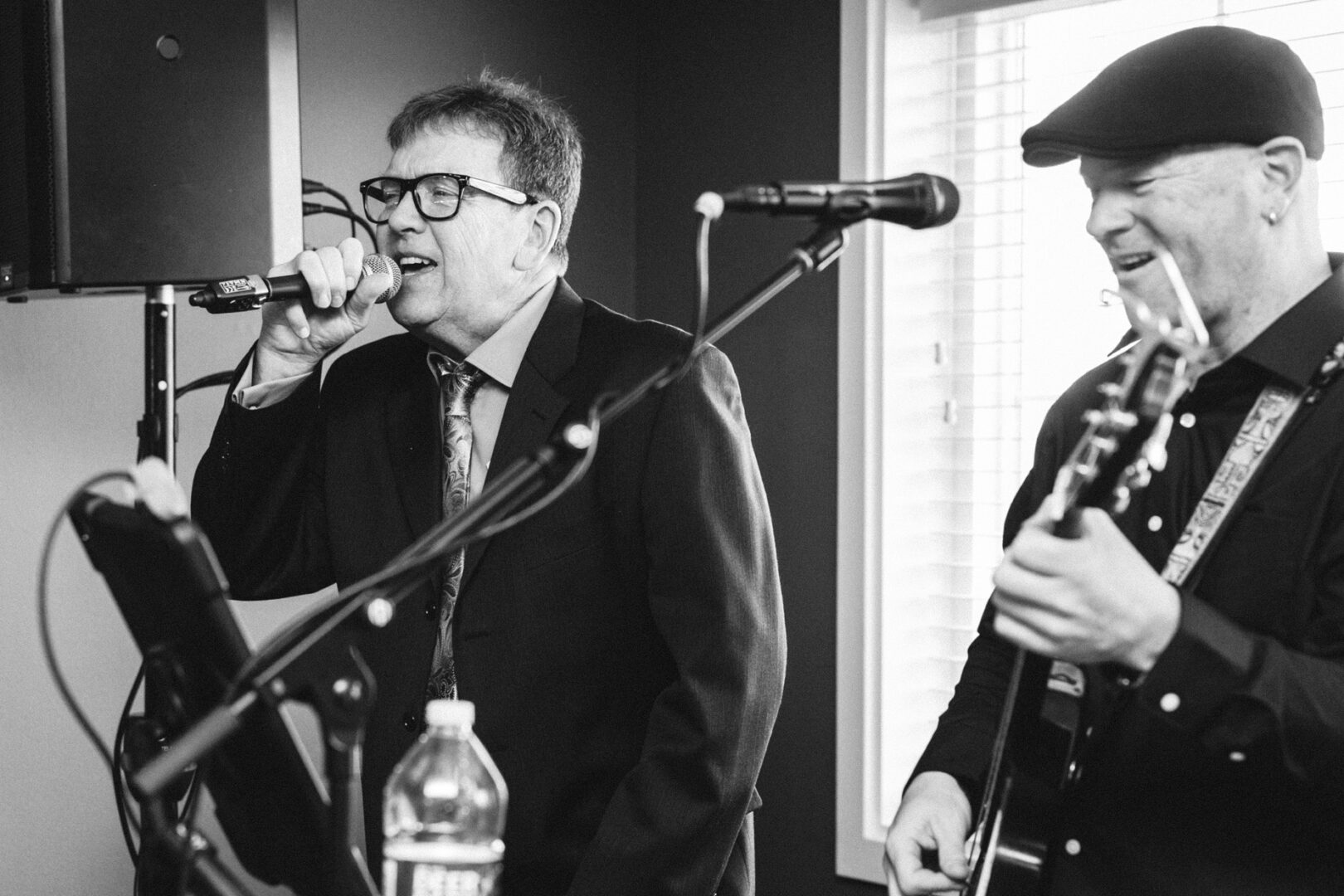 In the refined setting of an indoor wedding at the Yacht Club of Sea Isle City, two musicians captivate the audience; one sings soulfully into a microphone while the other masterfully plays an electric guitar. Both are elegantly dressed in suits, standing harmoniously before a grand window.
