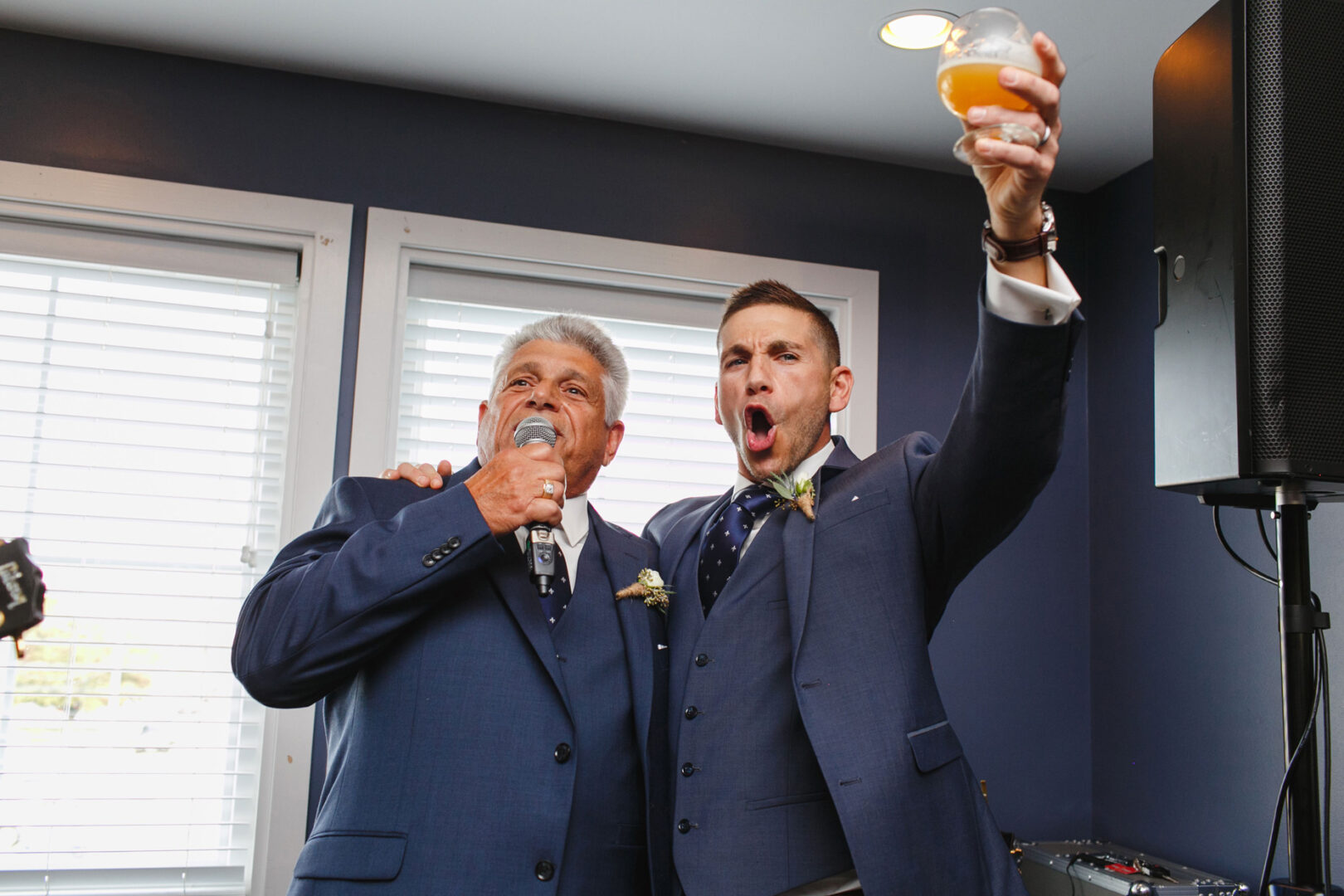 In a room with dark blue walls and white blinds, two men in blue suits stand together at the Yacht Club of Sea Isle City wedding, one holding a microphone while the other raises a glass.