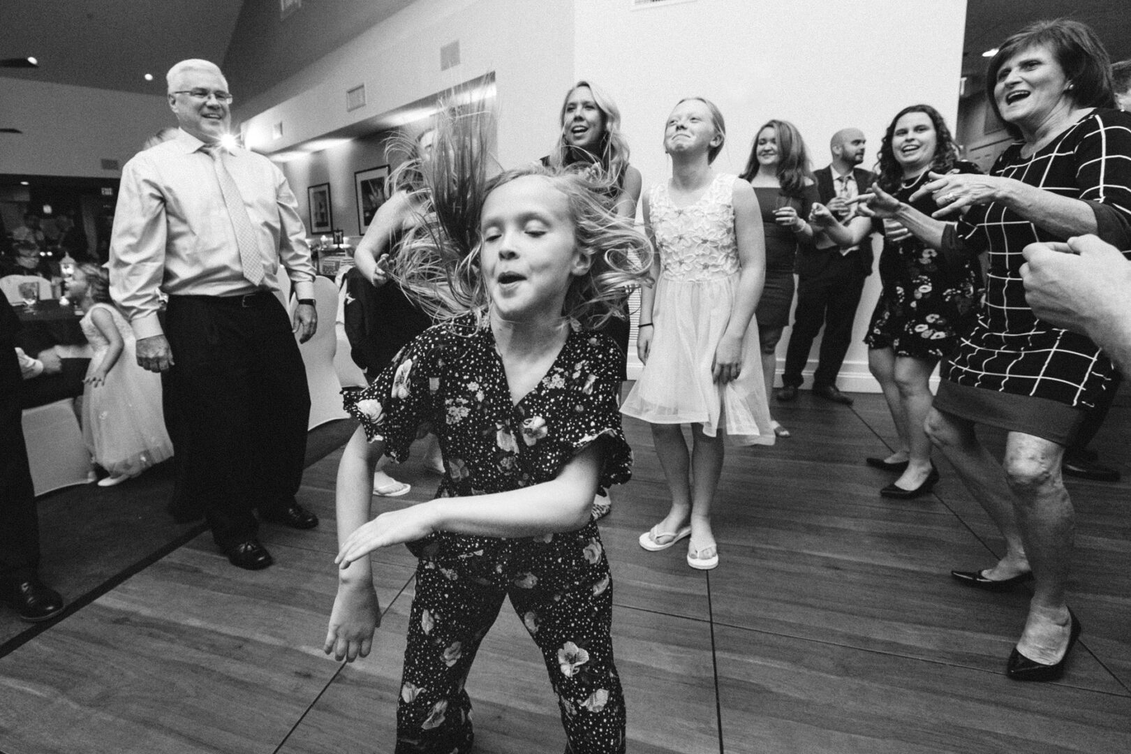 At a bustling Yacht Club of Sea Isle City wedding, a young girl energetically dances on the wooden floor as a delighted group of adults and children watch and smile in the background. The joyous scene unfolds indoors at this charming event.